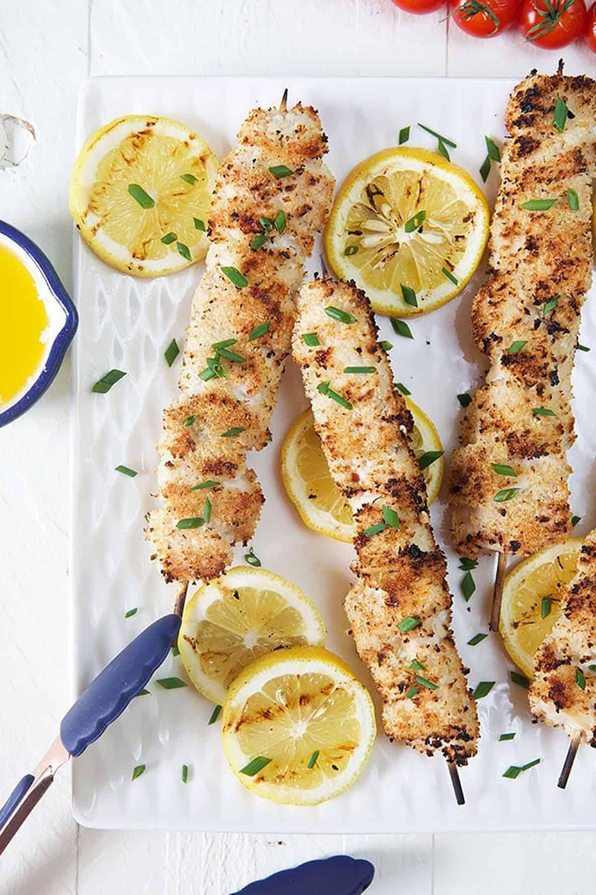 Spiedini di Pollo alla Griglia 2 Spiedini di pollo grigliati su un piatto bianco con limoni e pinze blu