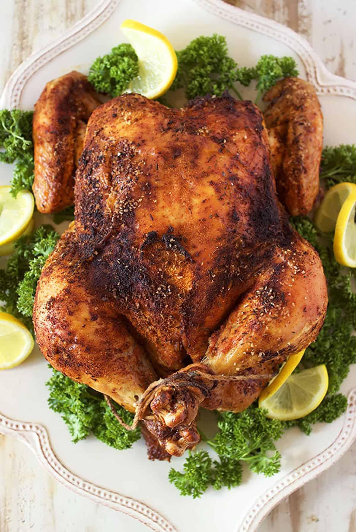 Come preparare il Pollo allo Spiedo 2 Un pollo arrosto intero su un piatto bianco con guarnizioni di prezzemolo e fette di limone.