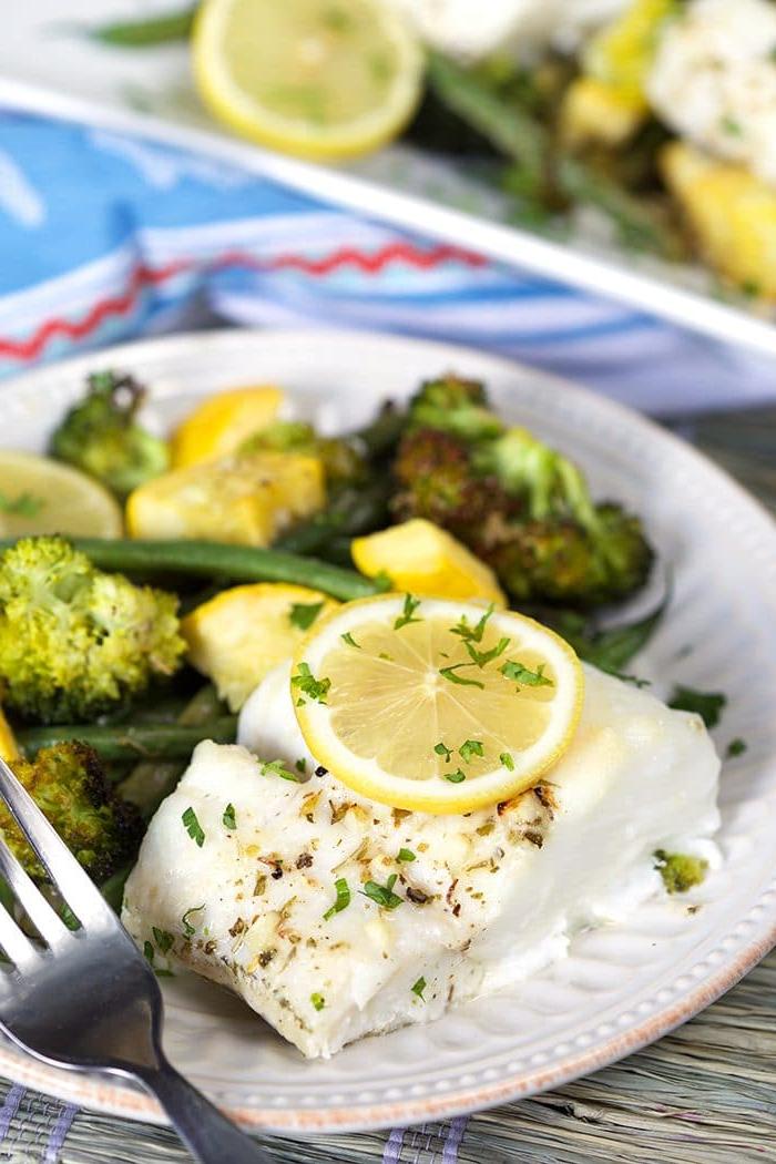 Merluzzo al Forno al Limone e Verdure 2 Merluzzo al forno al limone con una fetta di limone sopra e un mucchio di verdure verdi su un piatto bianco con una forchetta.