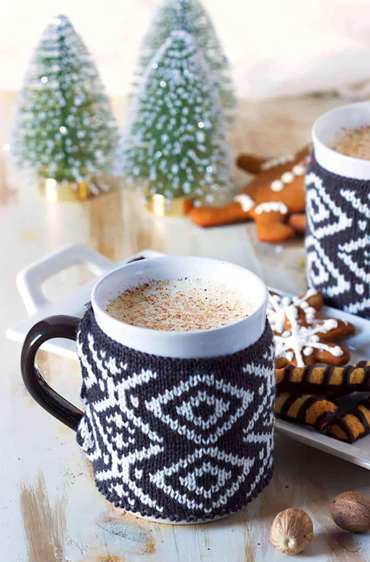La Migliore Ricetta Fatta in Casa di Zabaione 2 Tazza con una decorazione a maglia, piena di eggnog con due alberelli decorativi sullo sfondo.