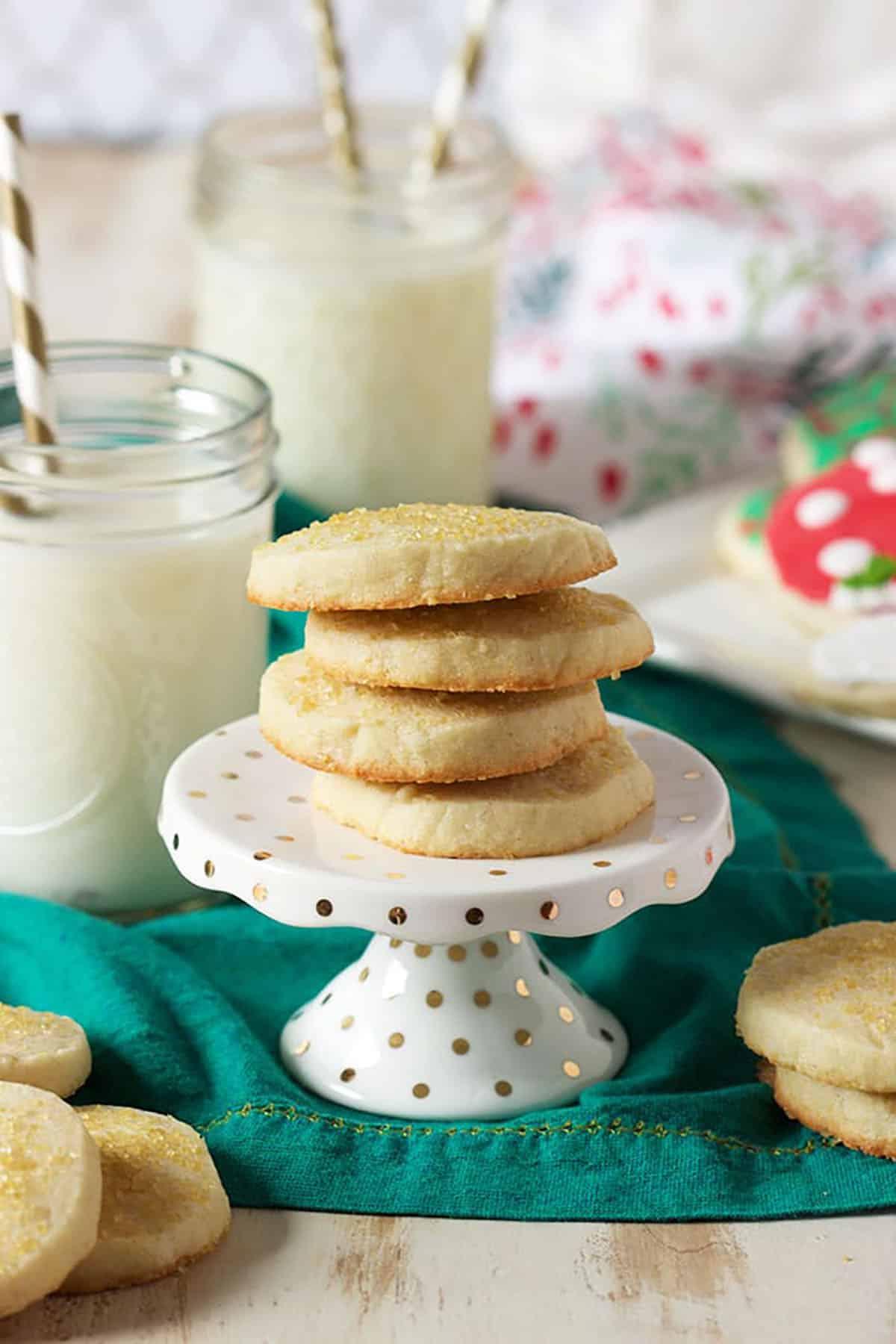 Biscotti di Natale allo zucchero 3 Biscotti di zucchero da affettare e cuocere su un'alzata per cupcake, su un tovagliolo verde con bicchieri di latte sullo sfondo. I biscotti di zucchero sono impilati uno sopra l'altro, pronti per essere gustati.