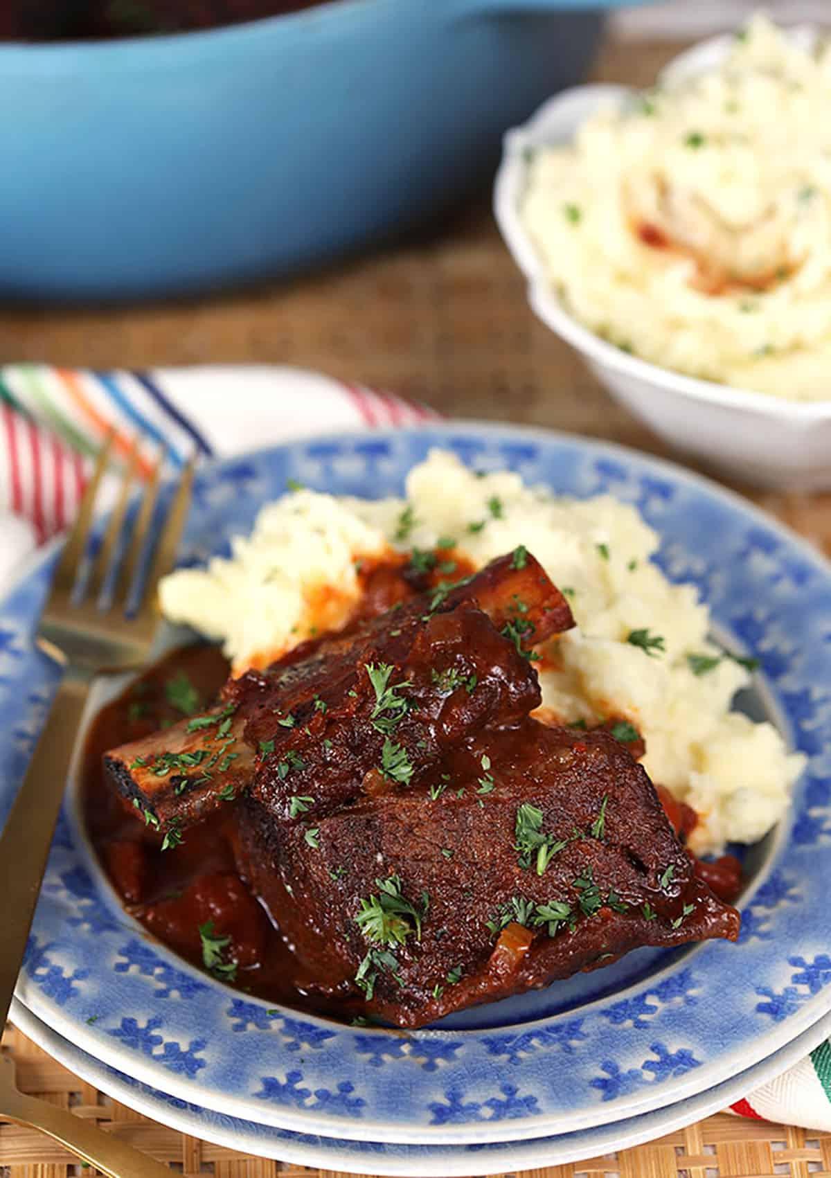 Brasato di Costine di Manzo al Miele e Chipotle 3 Costine di Manzo Brasate al Miele e Chipotle e Purè di Patate su un piatto blu con forchetta dorata.