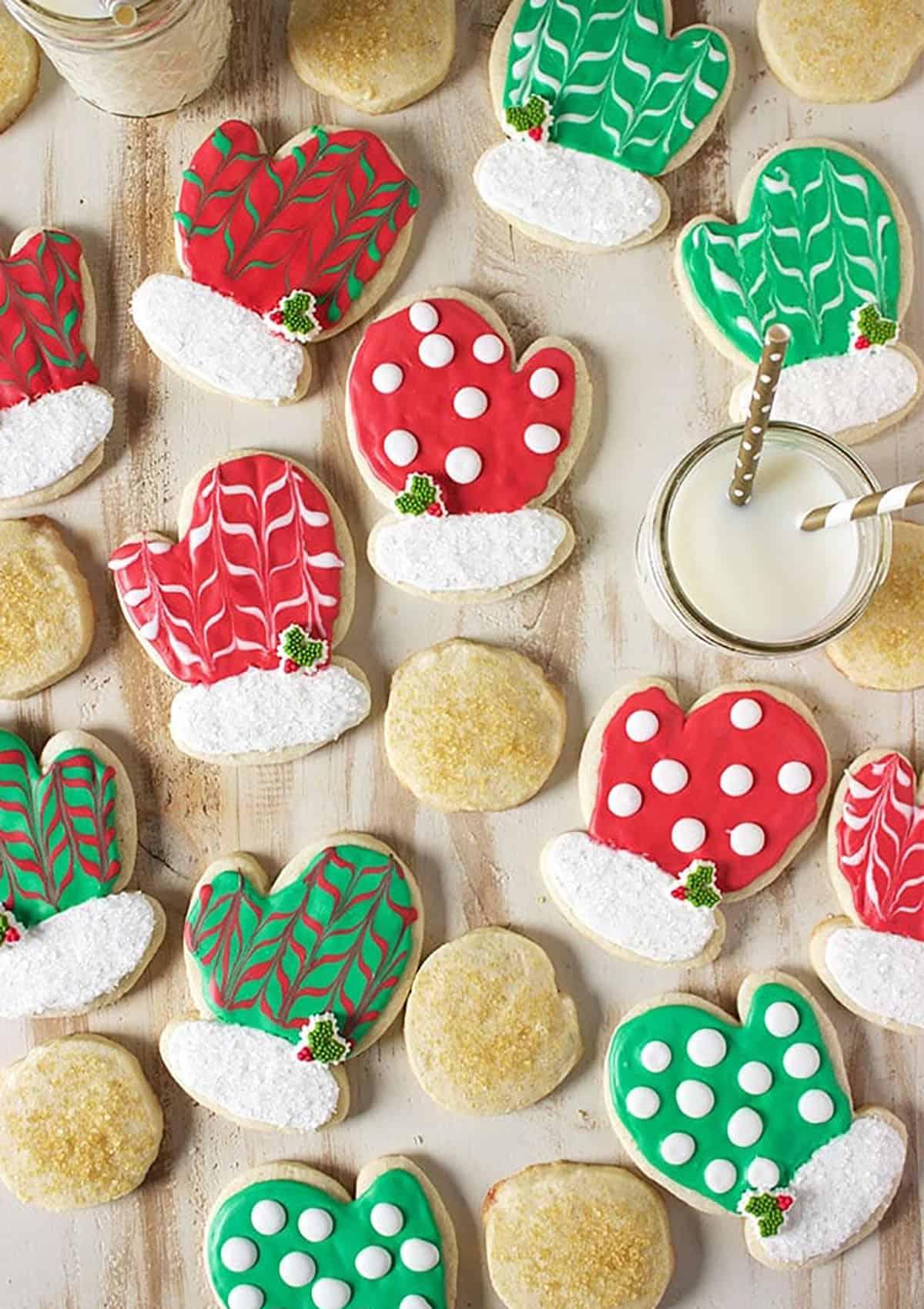 Biscotti di Natale allo zucchero 4 Biscotti di zucchero a forma di guanto decorati con glassa reale rossa e verde sparsi su una superficie di legno piatta, pronti per essere confezionati o gustati.