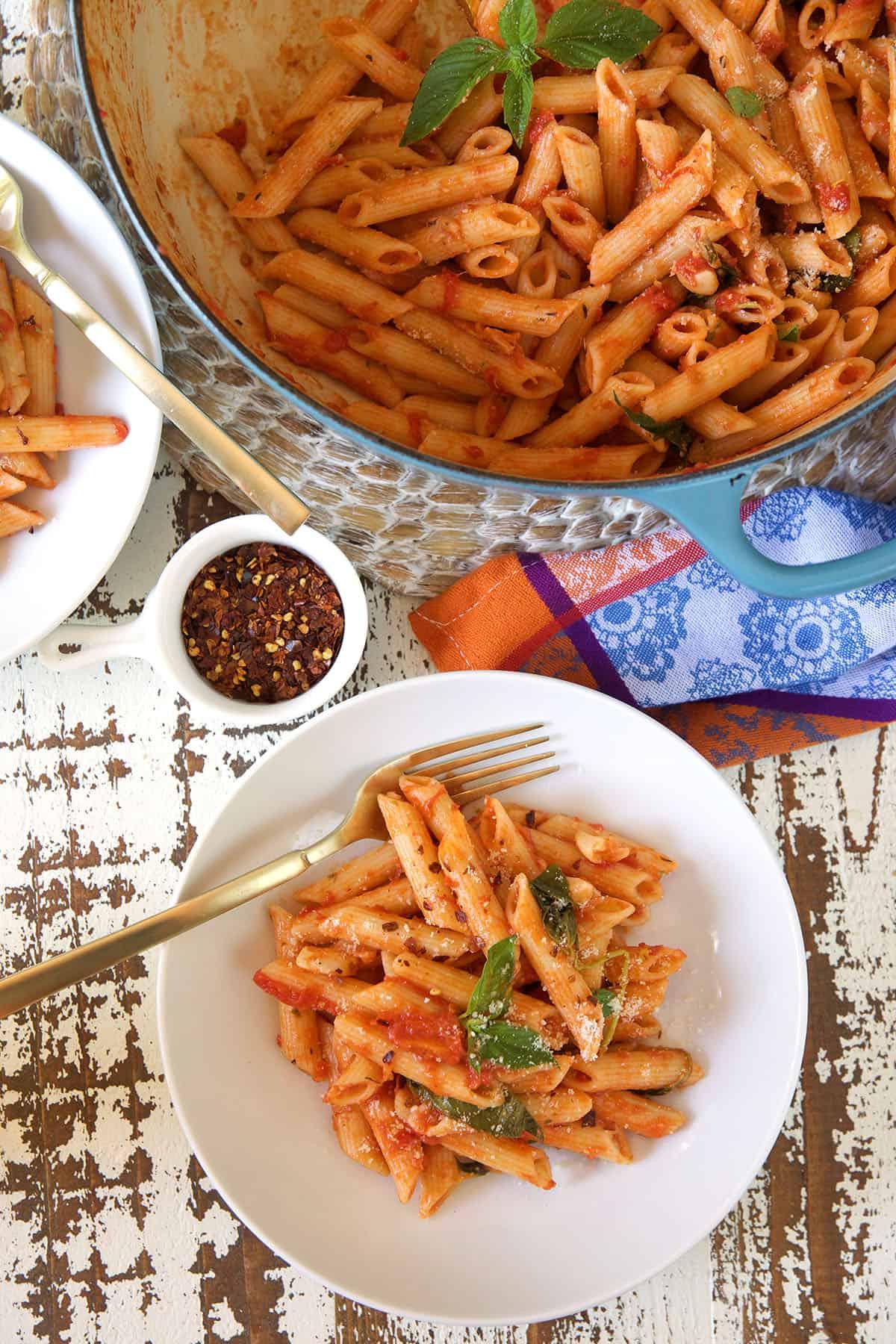 Penne al Pomodoro Veloce e Semplice 4 Un piatto bianco con una porzione di pasta è posizionato accanto a una pentola in ghisa piena e una piccola ciotola bianca con fiocchi di peperoncino rosso.