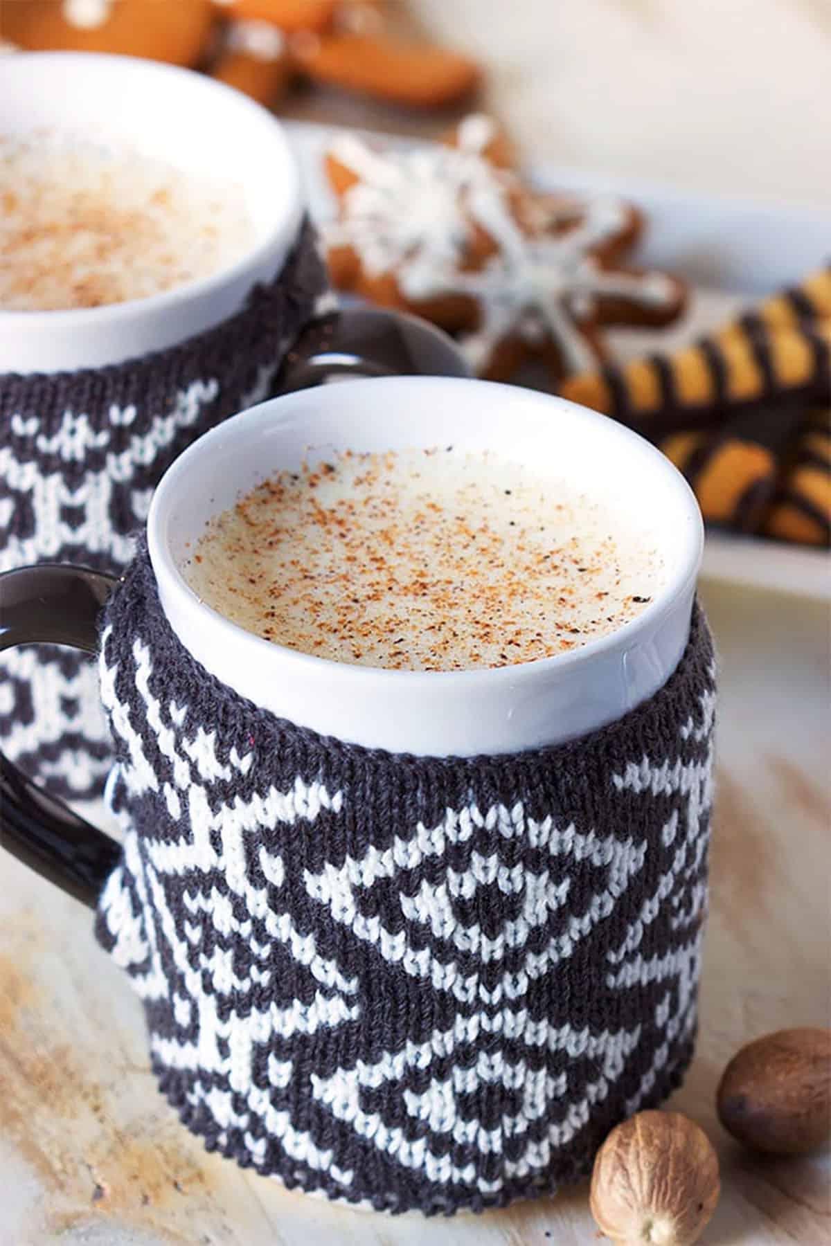 La Migliore Ricetta Fatta in Casa di Zabaione 4 Due tazze con decorazioni a maglia riempite di eggnog con biscotti di pan di zenzero sullo sfondo.