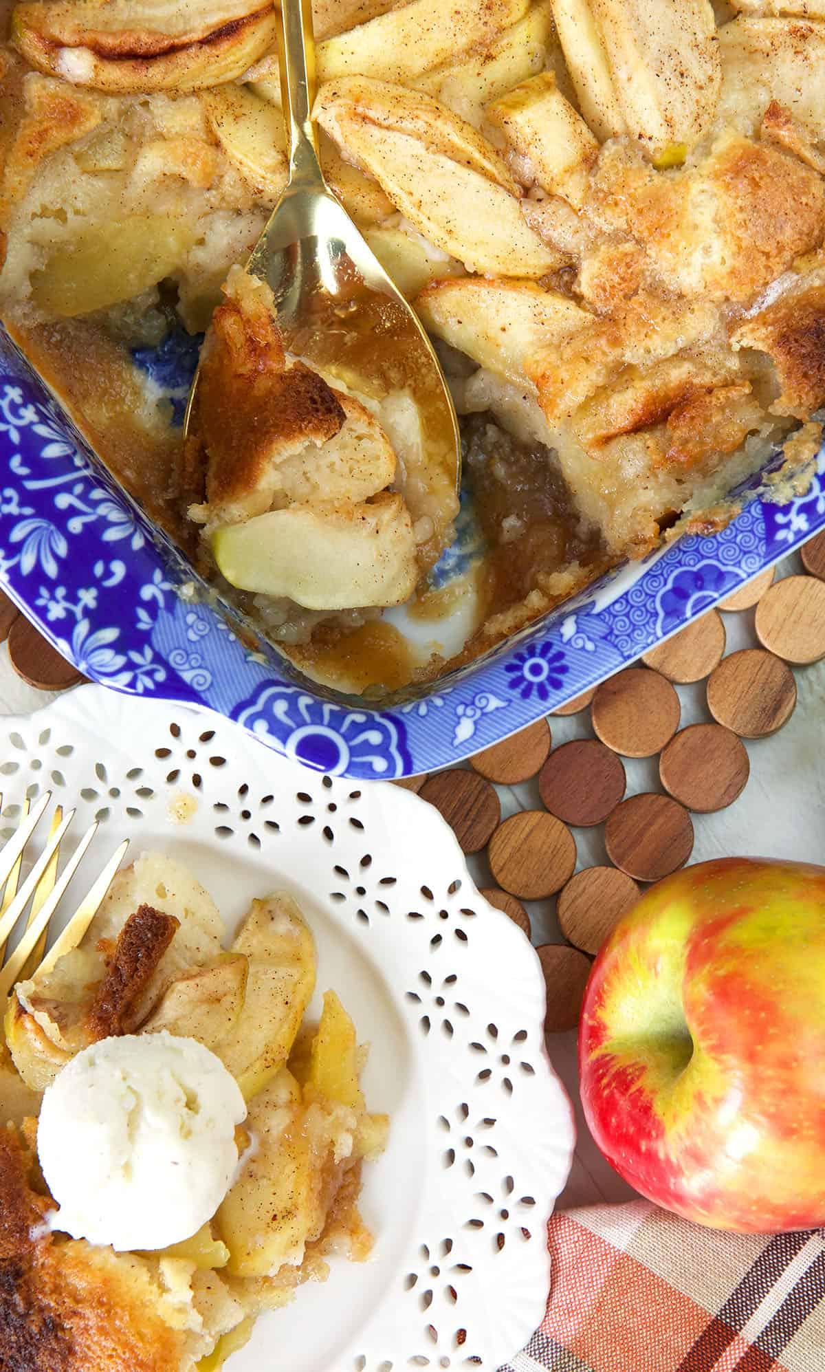 Una singola porzione di cobbler di mele, guarnita con gelato, è posta accanto a un cobbler intero nella pirofila blu.
