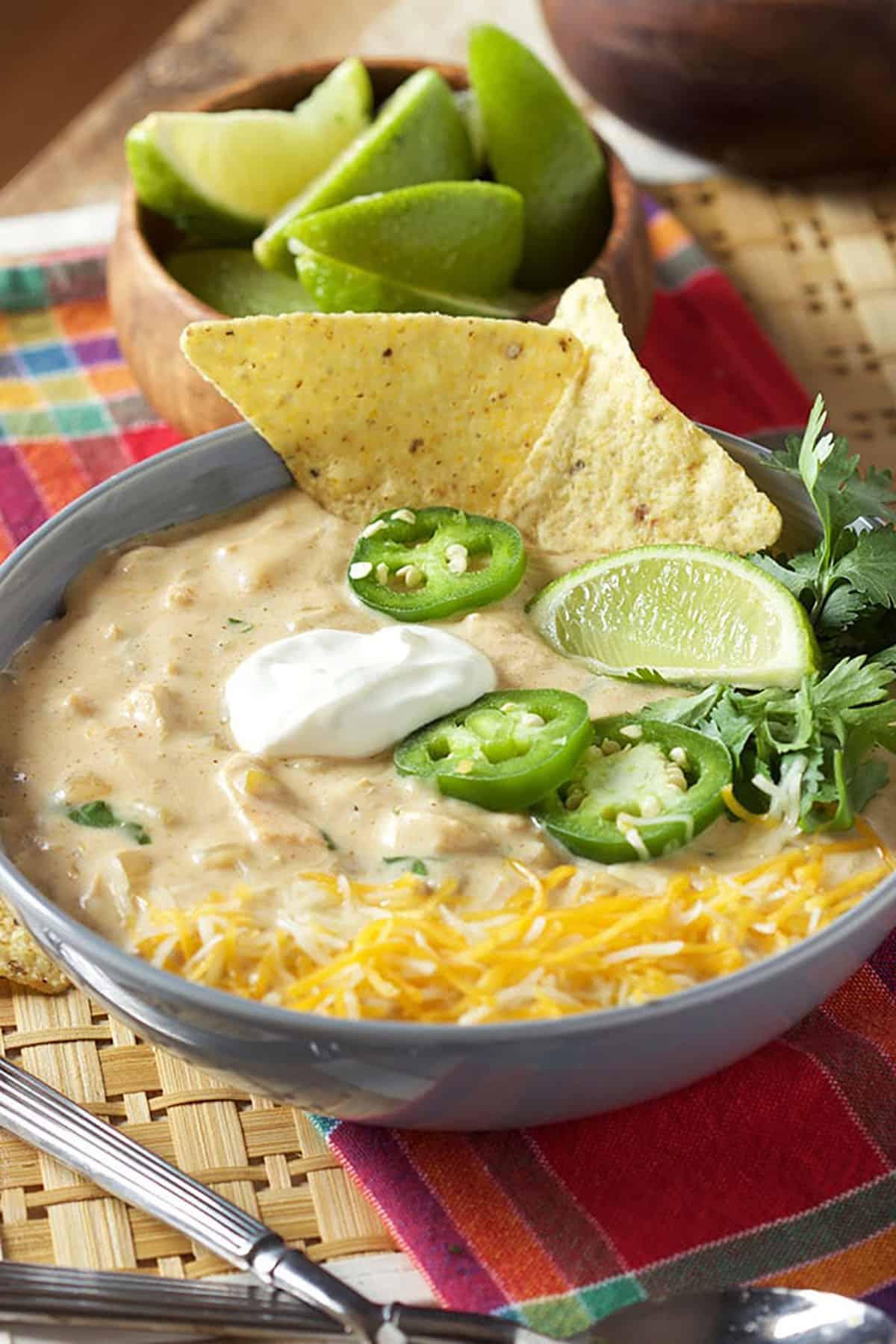 Il Miglior Chili Bianco Cremoso di Pollo 5 Chili bianco di pollo in una ciotola grigia con patatine e jalapeño sopra. Ciotola di lime sullo sfondo.