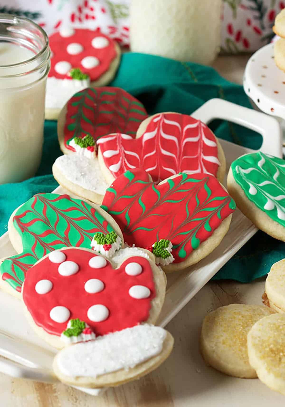 Biscotti di Natale allo zucchero 5 Biscotti di zucchero a forma di guanto decorati con glassa reale rossa e verde su un piatto bianco con un bicchiere di latte fresco sullo sfondo.