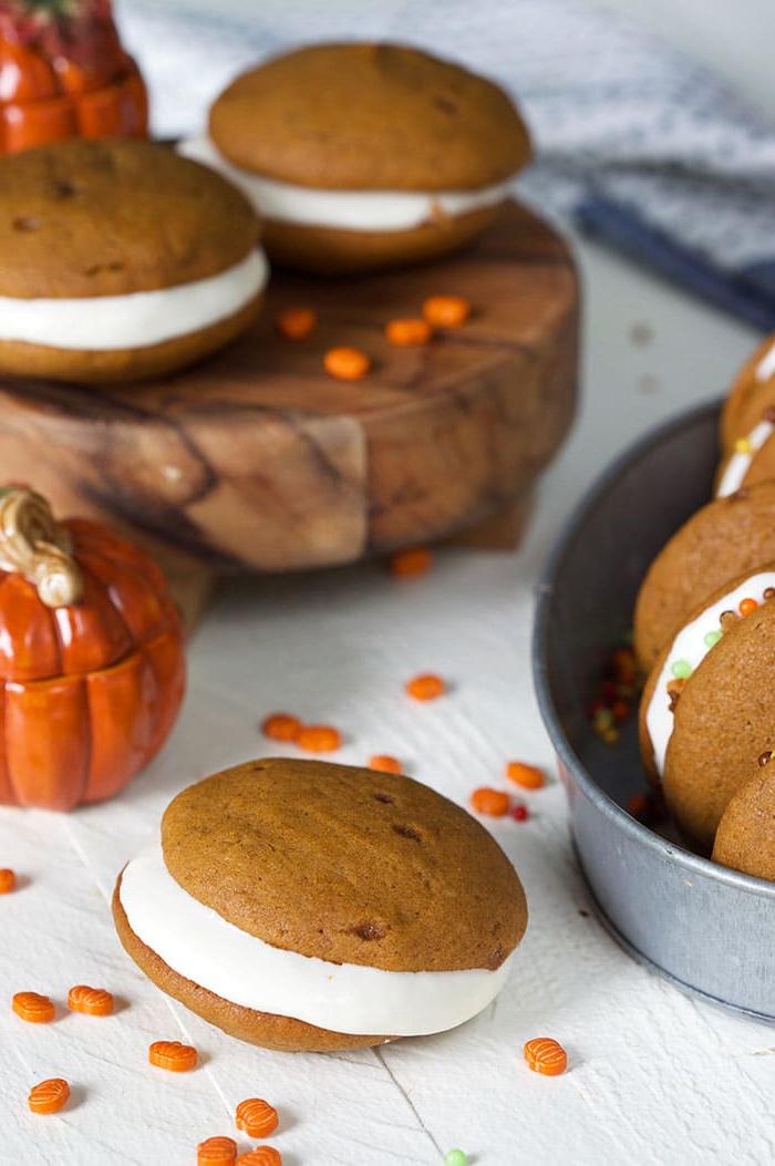 **Whoopie Pies alla Zucca con Ripieno al Mascarpone d'Acero** 6 Whoopie pies alla zucca su un ceppo di legno con una zucca di ceramica davanti.