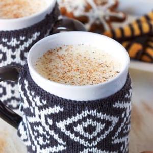 La Migliore Ricetta Fatta in Casa di Zabaione 7 Due tazze con decorazioni a maglia riempite di eggnog con biscotti di pan di zenzero sullo sfondo.