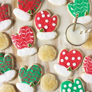 Biscotti di Natale allo zucchero 8 Biscotti di zucchero a forma di guanto decorati con glassa reale rossa e verde sparsi su una superficie di legno piatta.