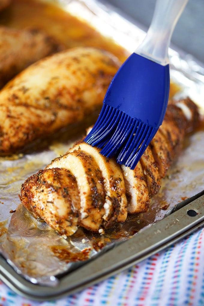 Le migliori ricette di petto di pollo al forno 9 Petto di pollo al forno a fette che viene spennellato con un pennello di silicone blu su una teglia foderata di carta stagnola.