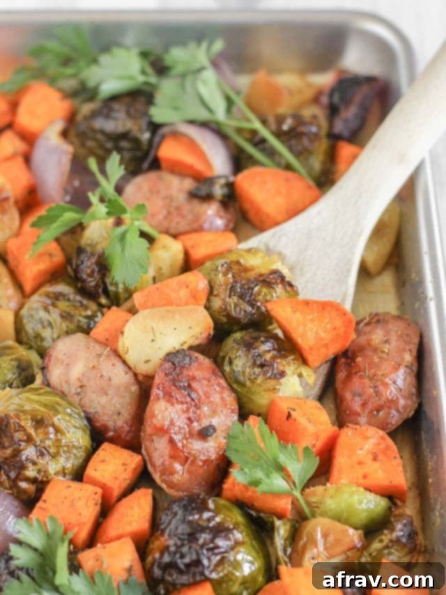 Roasted vegetables and chicken sausage on a large baking sheet with a wooden spatula scooping up some of the food.