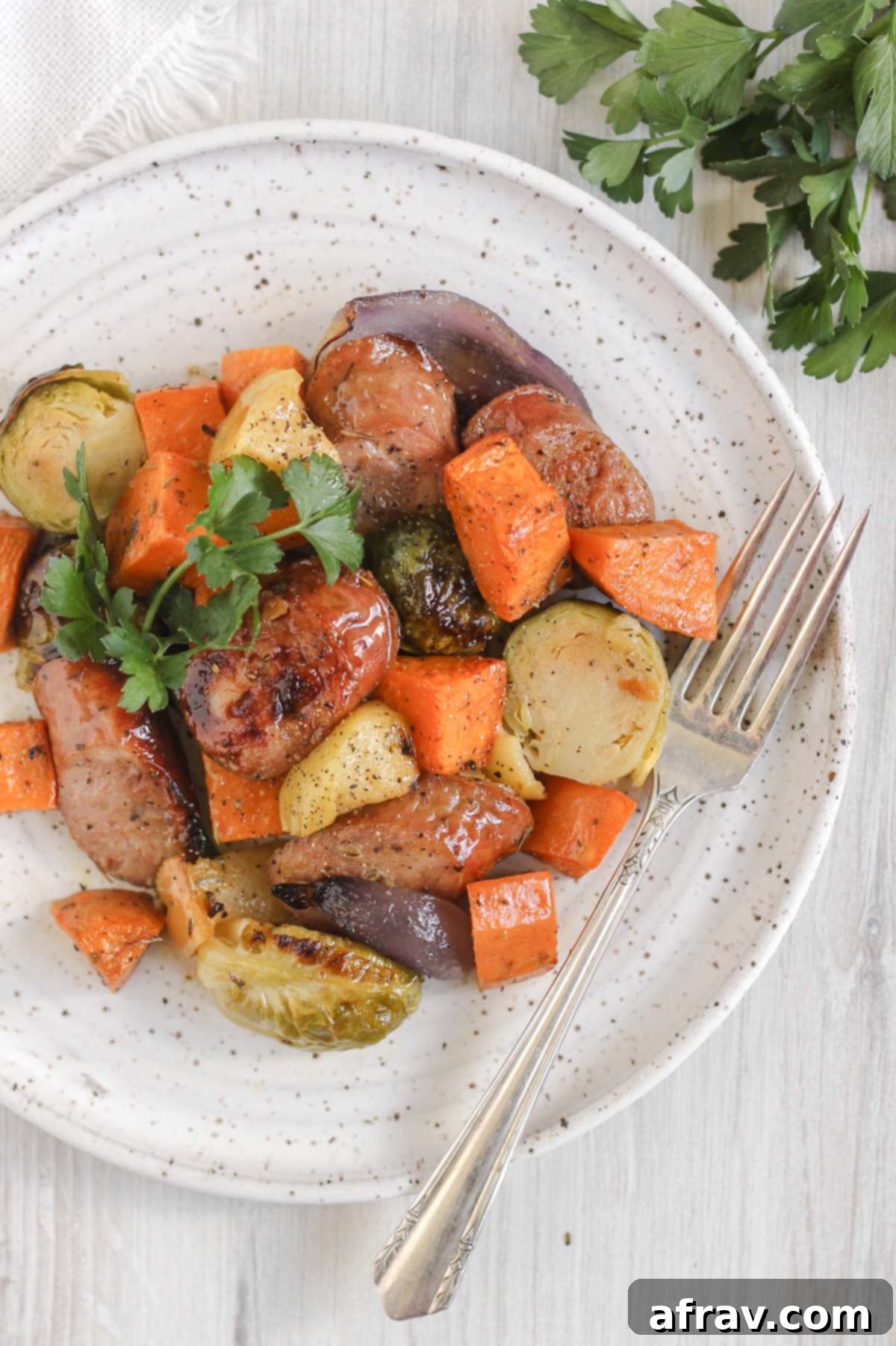 Chicken sausage and roasted veggies with a vintage fork on a round white plate with parsley off to the side.