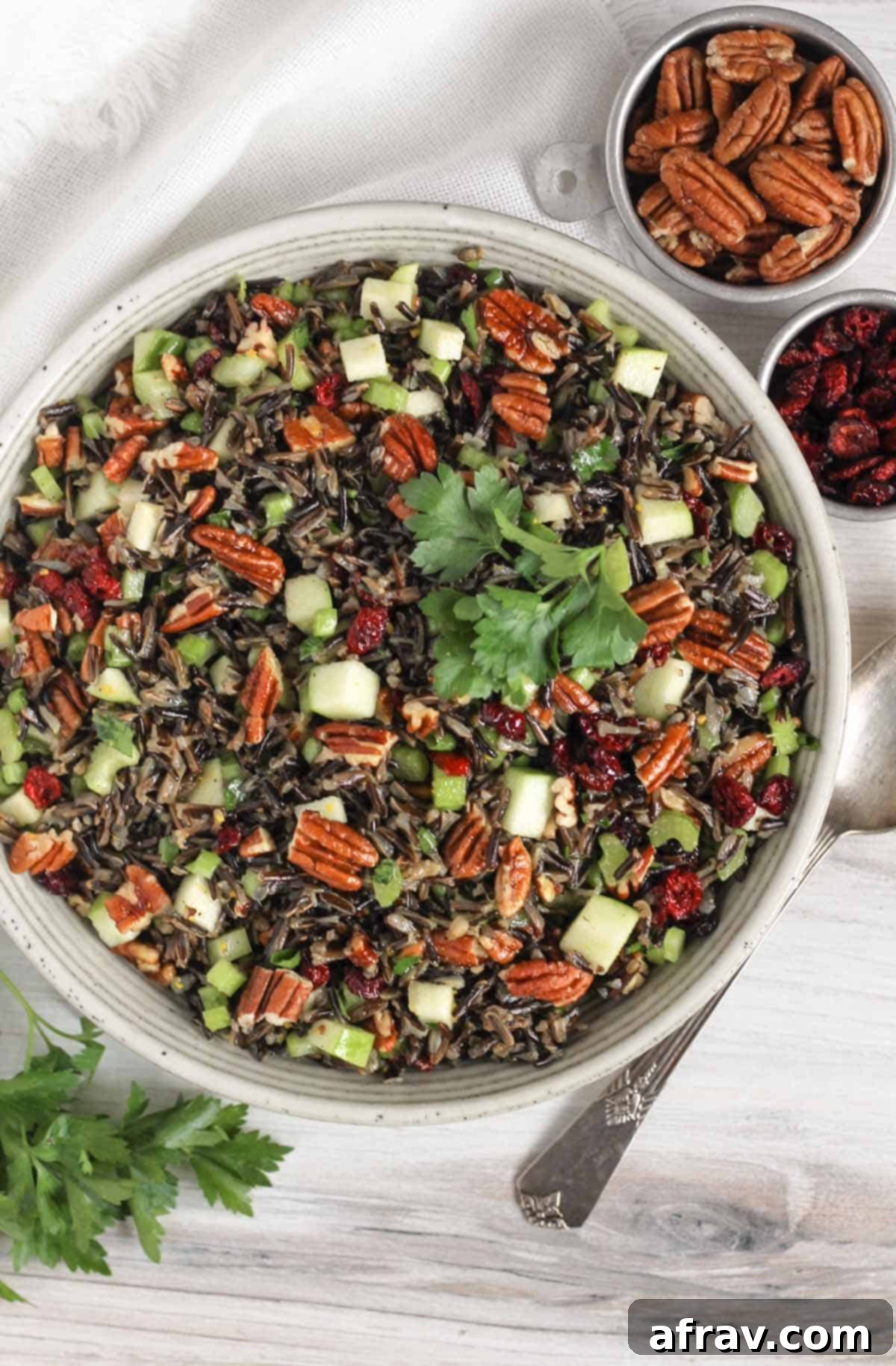 Wild rice salad in a ceramic serving bowl with a vintage serving spoon in the salad and a small dish of dried cranberries and pecans on the side.