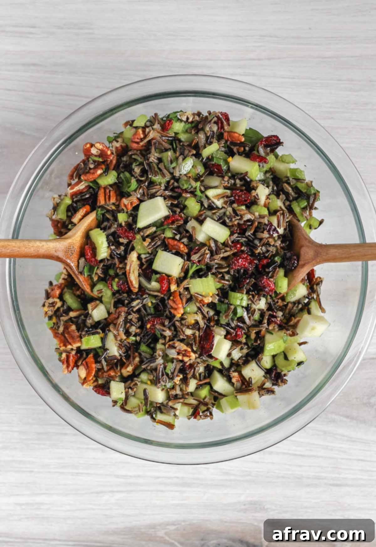 Two wooden salad utensils mixing wild rice salad ingredients in a glass mixing bowl.