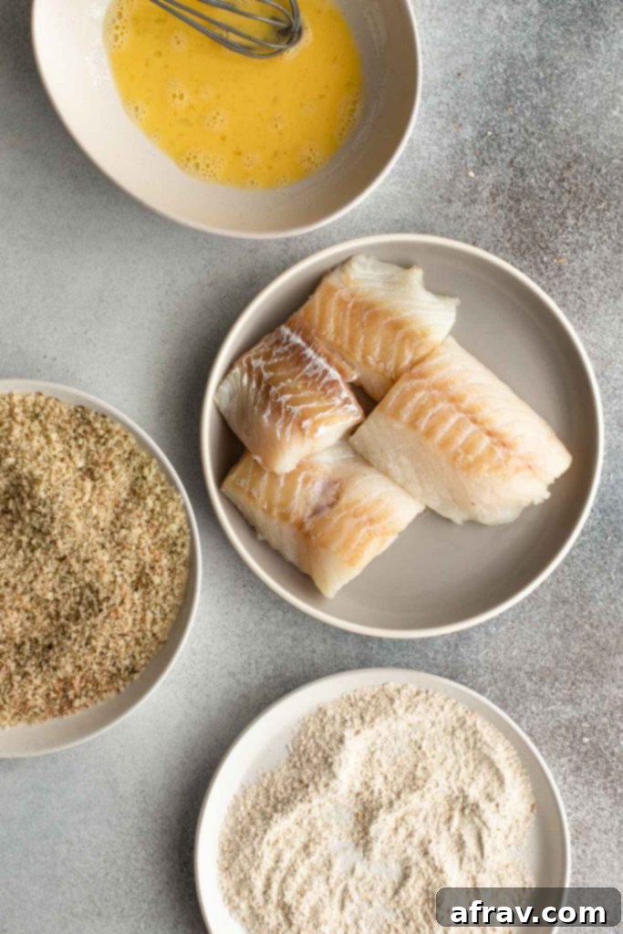 Overhead view of cod, scrambled egg, panko crumbs and flour arranged on separate plates/bowls.