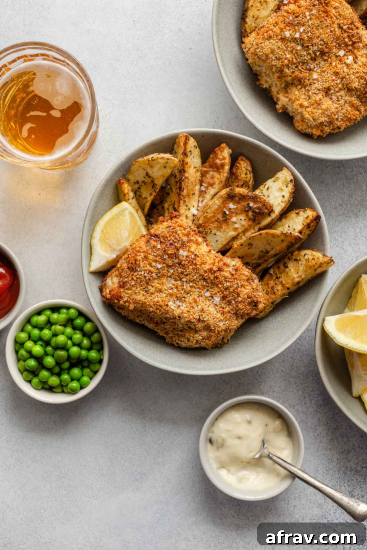 Overhead view of two bowls of breaded fish and wedge fries with peas and tartar sauce off to the sides.