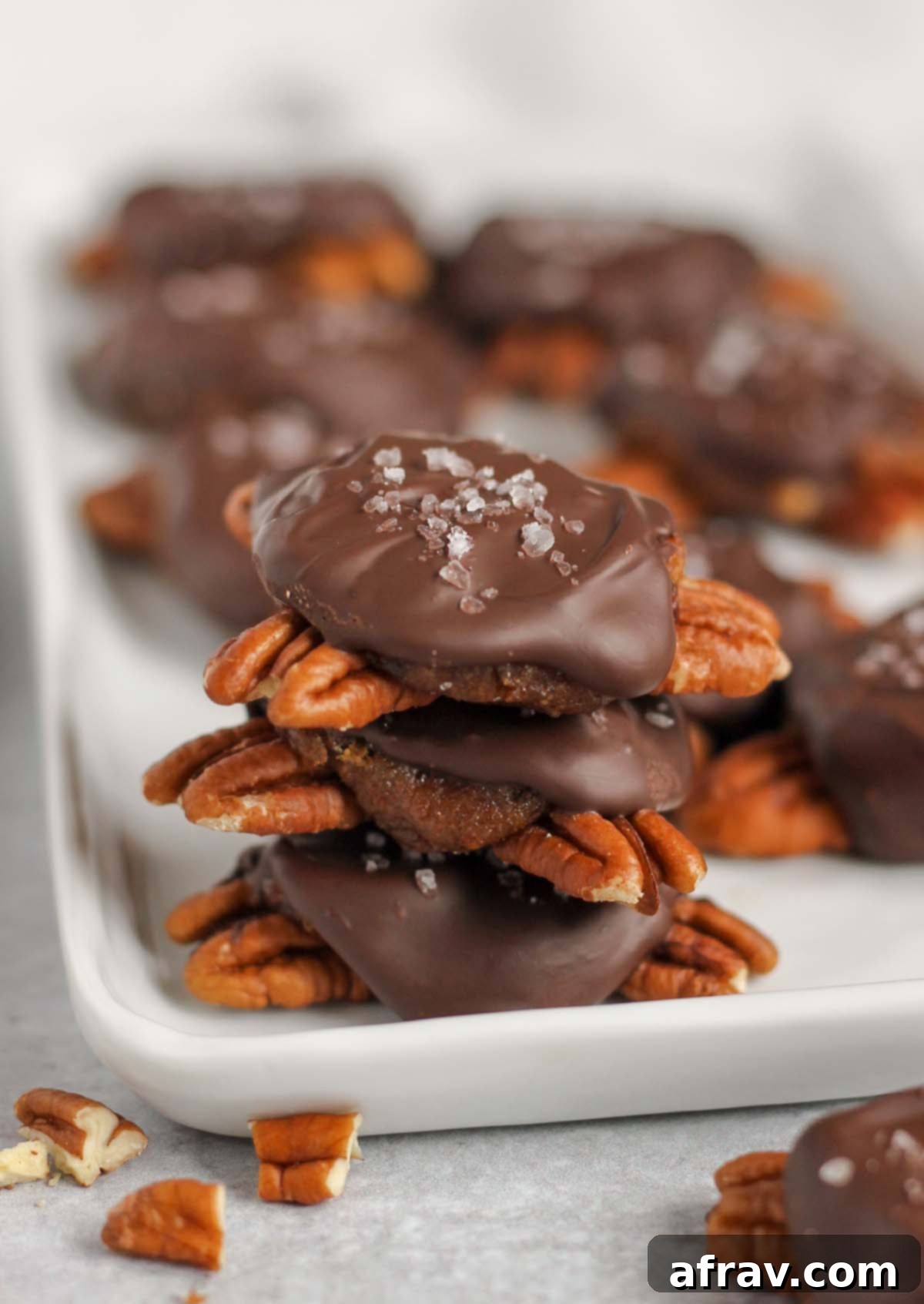 Chocolate turtle candies stacked 3high on a white serving tray and extra turtles in the background.