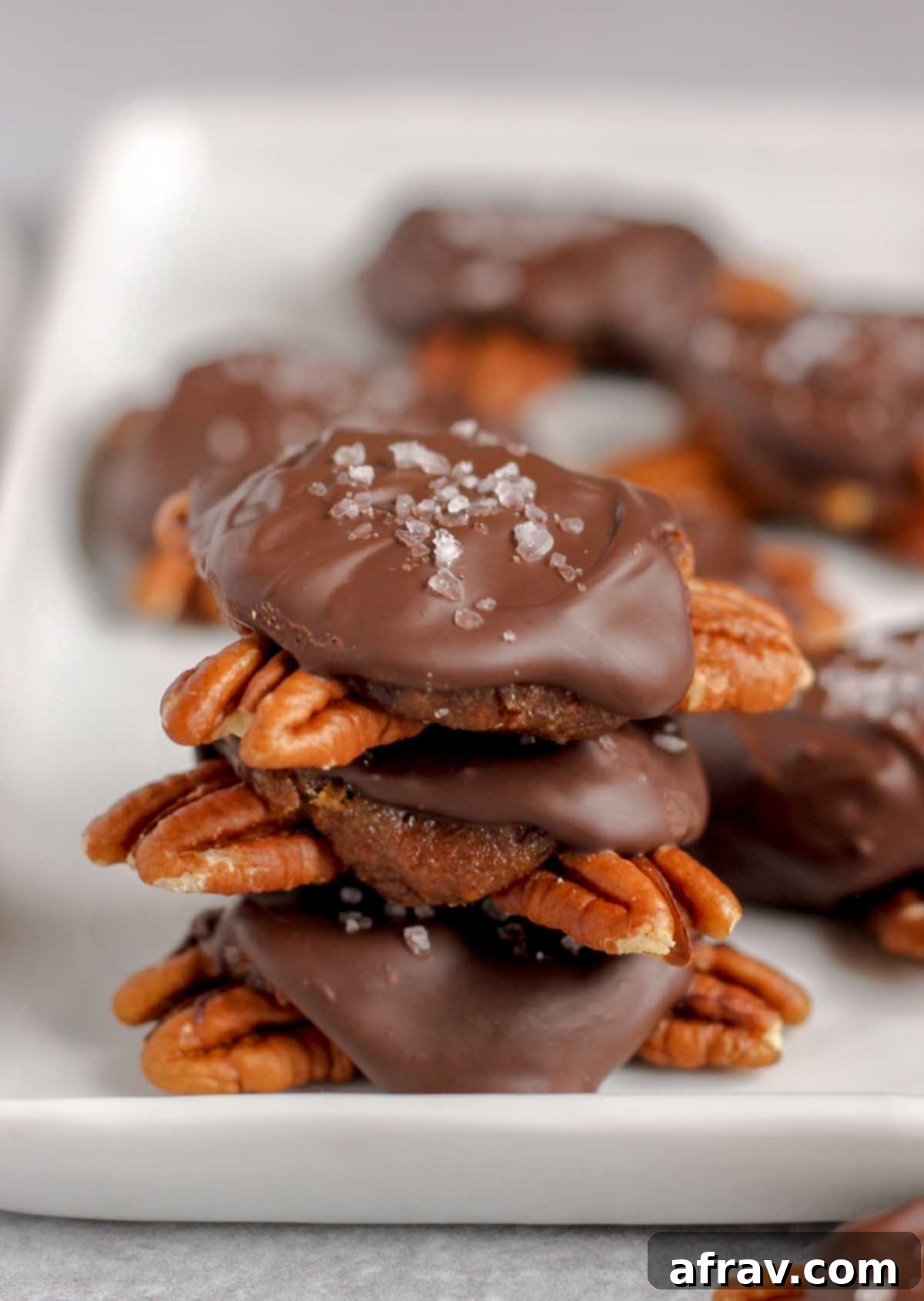 Chocolate turtle candies stacked 3high on a white serving tray and extra turtles in the background.