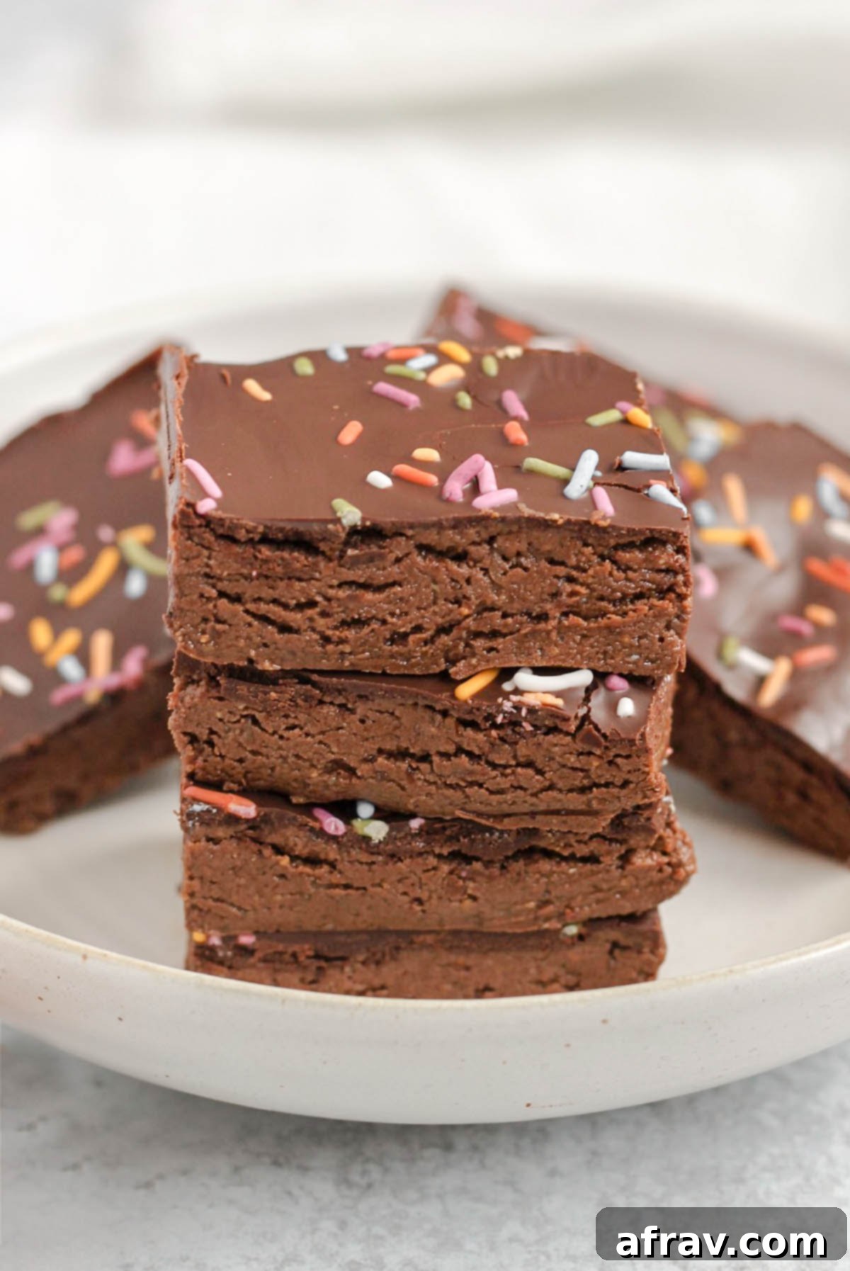 Homemade No-Bake Chocolate Protein Bars 5 Square pieces of chocolate protein bar stacked 4 high on a plate with other bars in the background.