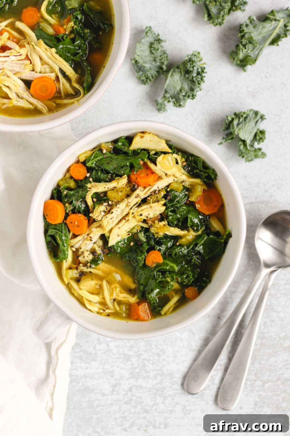 Chicken and kale soup in a white bowl with two soup spoons laid on the side.