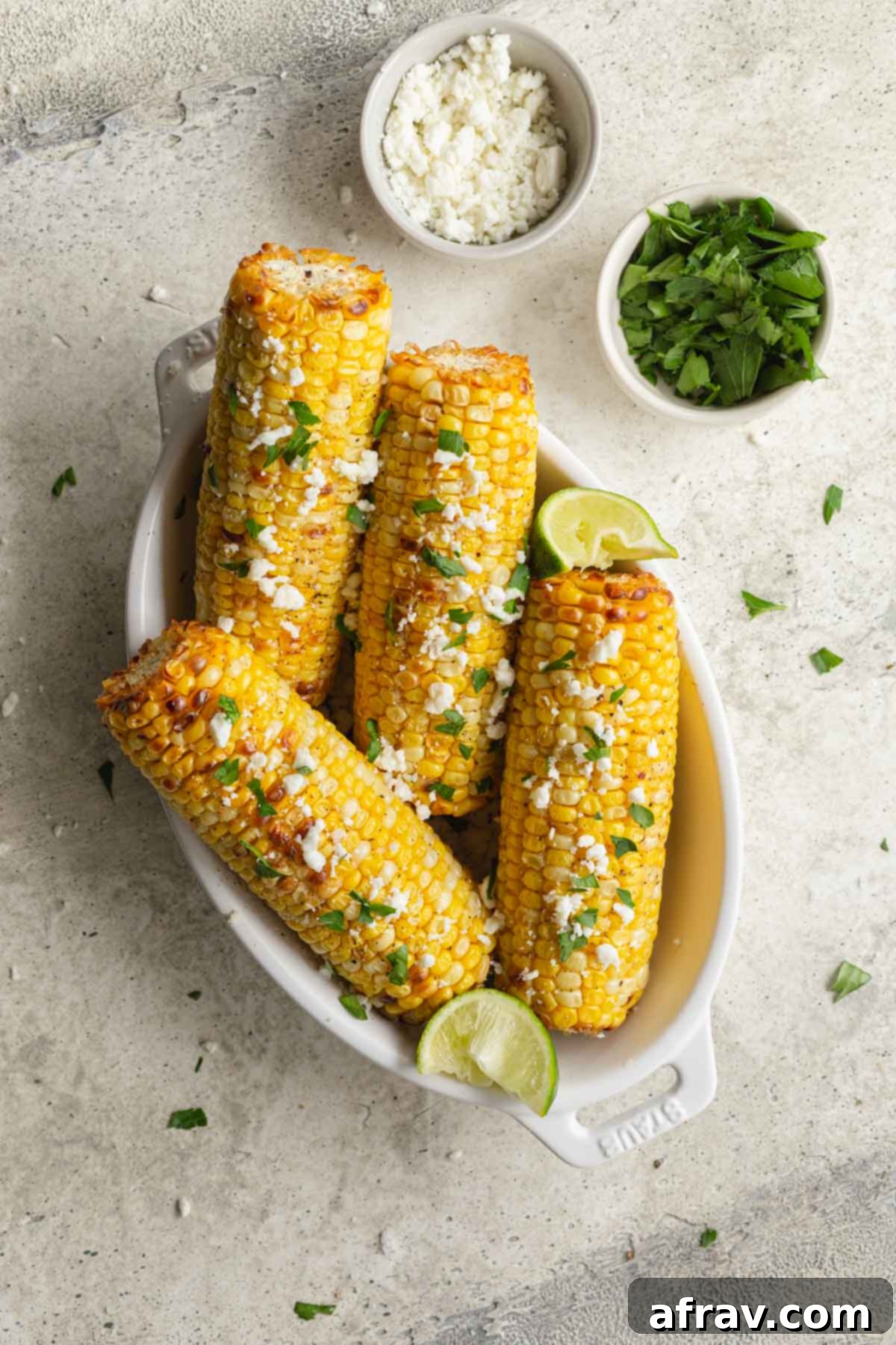 Overhead view of four ears of air fried corn in an oval dish with bowls of feta and chopped parsley off to the side. 