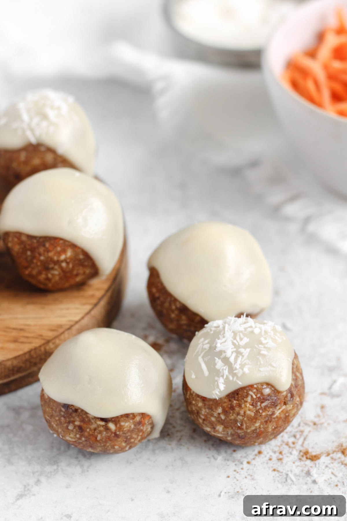 Three carrot cake bites topped with glaze on a gray surface with a bowl of shredded carrots in the background.