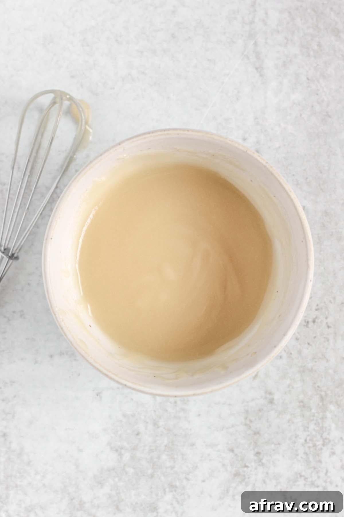 Coconut butter glaze in a small white bowl with a mini whisk dripping in glaze off to the side. 