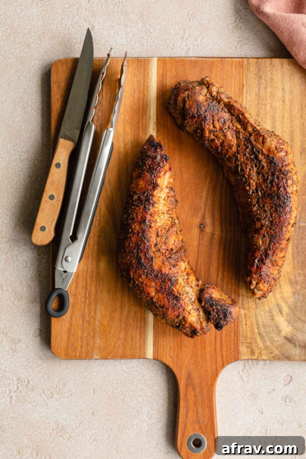Perfect Air Fryer Pork Tenderloin 8 Two perfectly cooked pork tenderloins on a wooden cutting board next to tongs and a knife, ready for slicing.