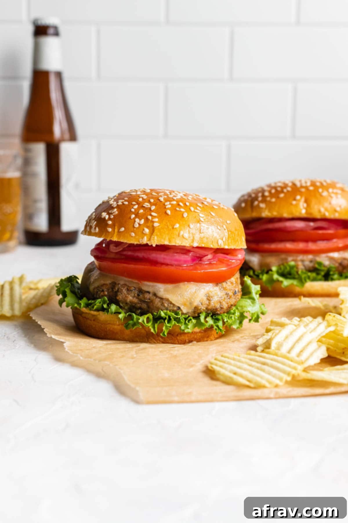Side view of two burgers on parchment paper with potato chips and a bottle of beer in the background.