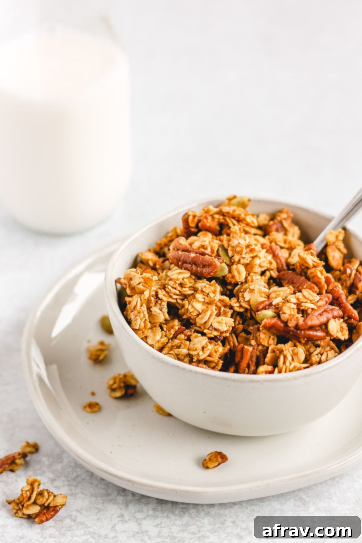 Wholesome Pumpkin Crunch Granola Gluten Free Plant Based 2 A bowl of homemade pumpkin granola on a small plate, with a glass jar of milk in the background. The granola is golden brown and chunky.