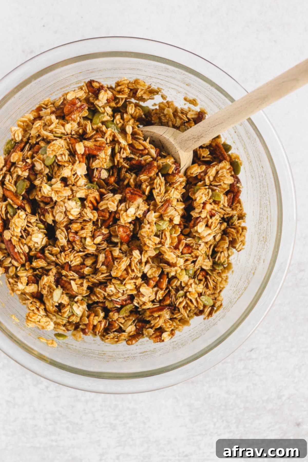 Wholesome Pumpkin Crunch Granola Gluten Free Plant Based 4 Pumpkin granola ingredients thoroughly mixed together in a large glass mixing bowl with a wooden spoon ready to be transferred to a baking sheet.