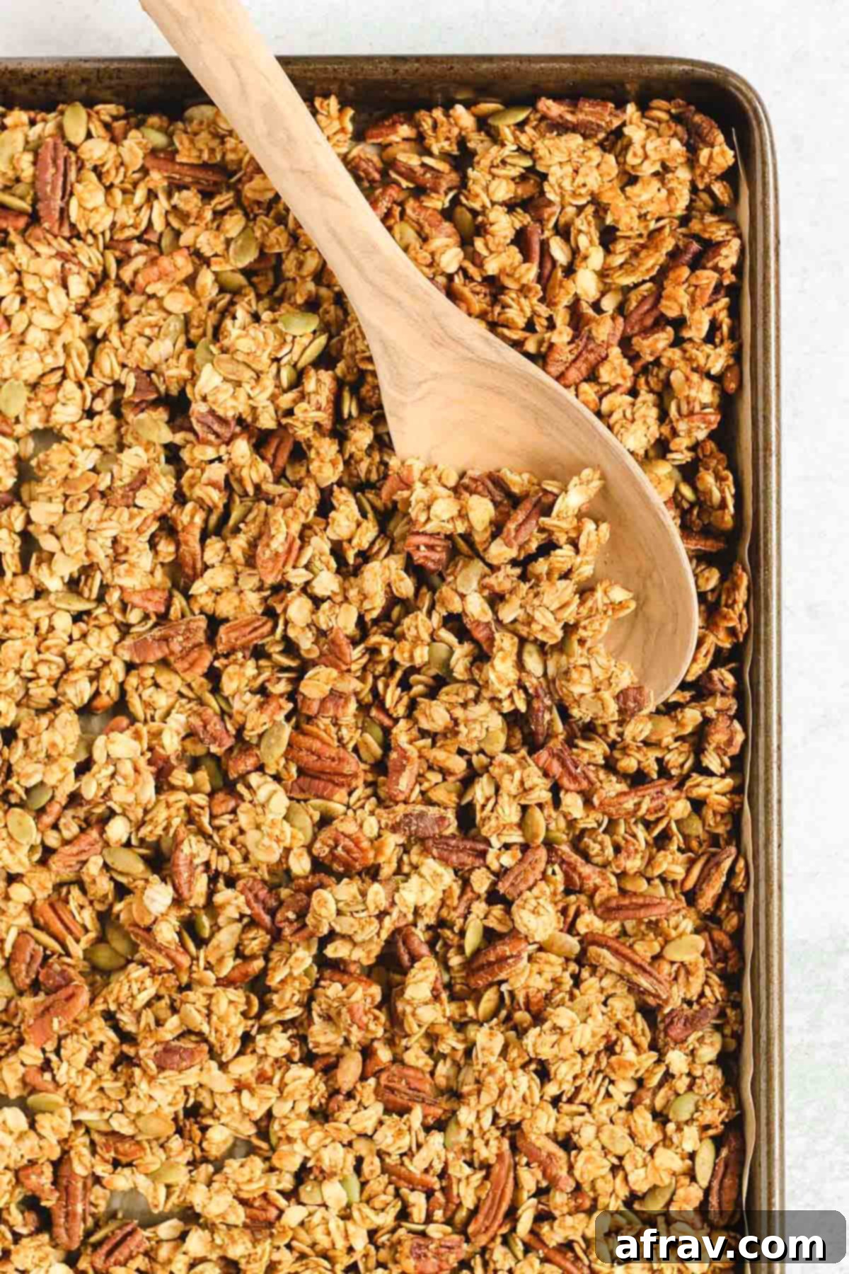 Wholesome Pumpkin Crunch Granola Gluten Free Plant Based 6 Freshly baked pumpkin granola on a large baking sheet, with a wooden spoon scooping up some of the crunchy clusters.