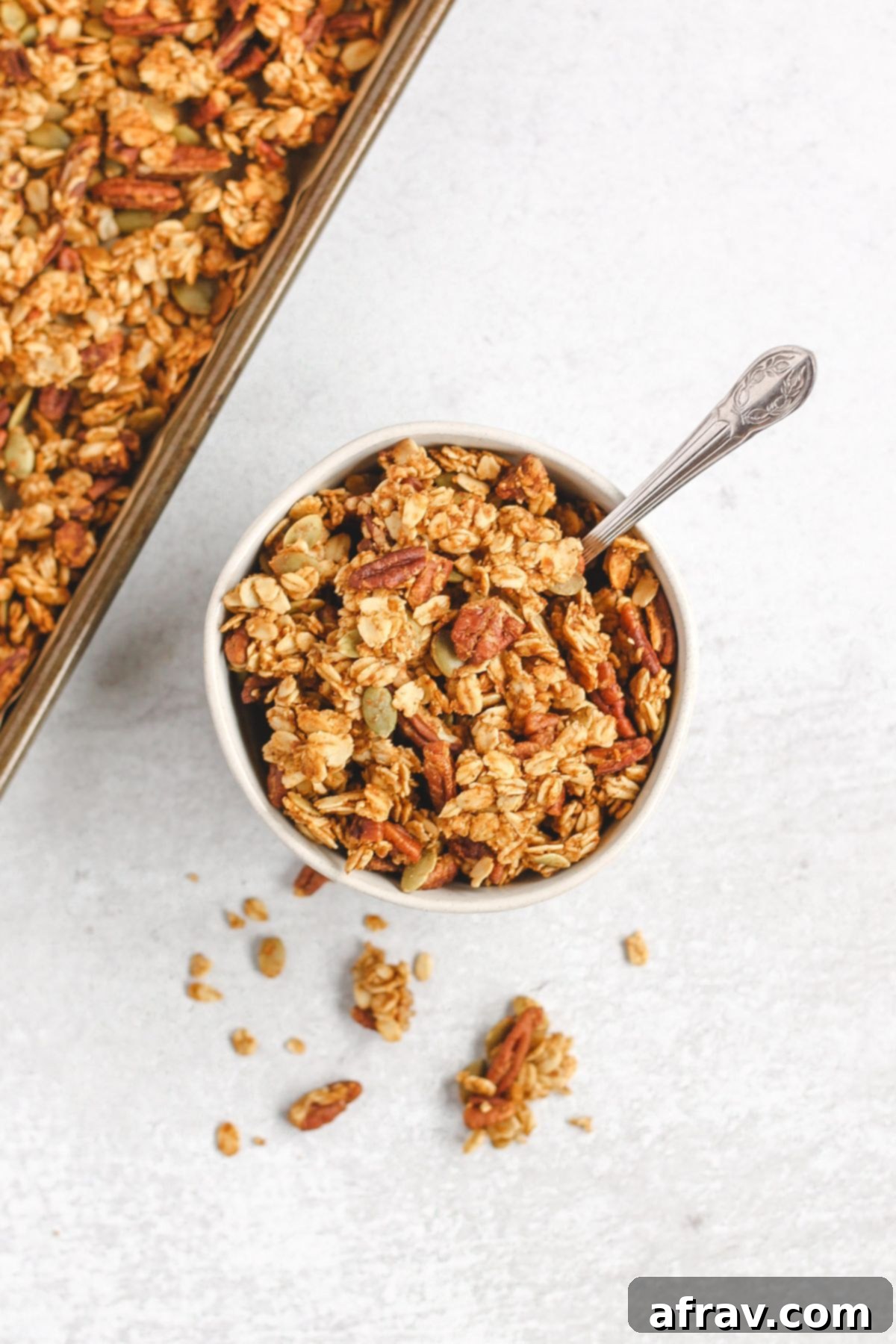 Wholesome Pumpkin Crunch Granola Gluten Free Plant Based 7 A small bowl of pumpkin granola with a vintage spoon, next to a baking sheet filled with more granola. It evokes a cozy, homemade feel.