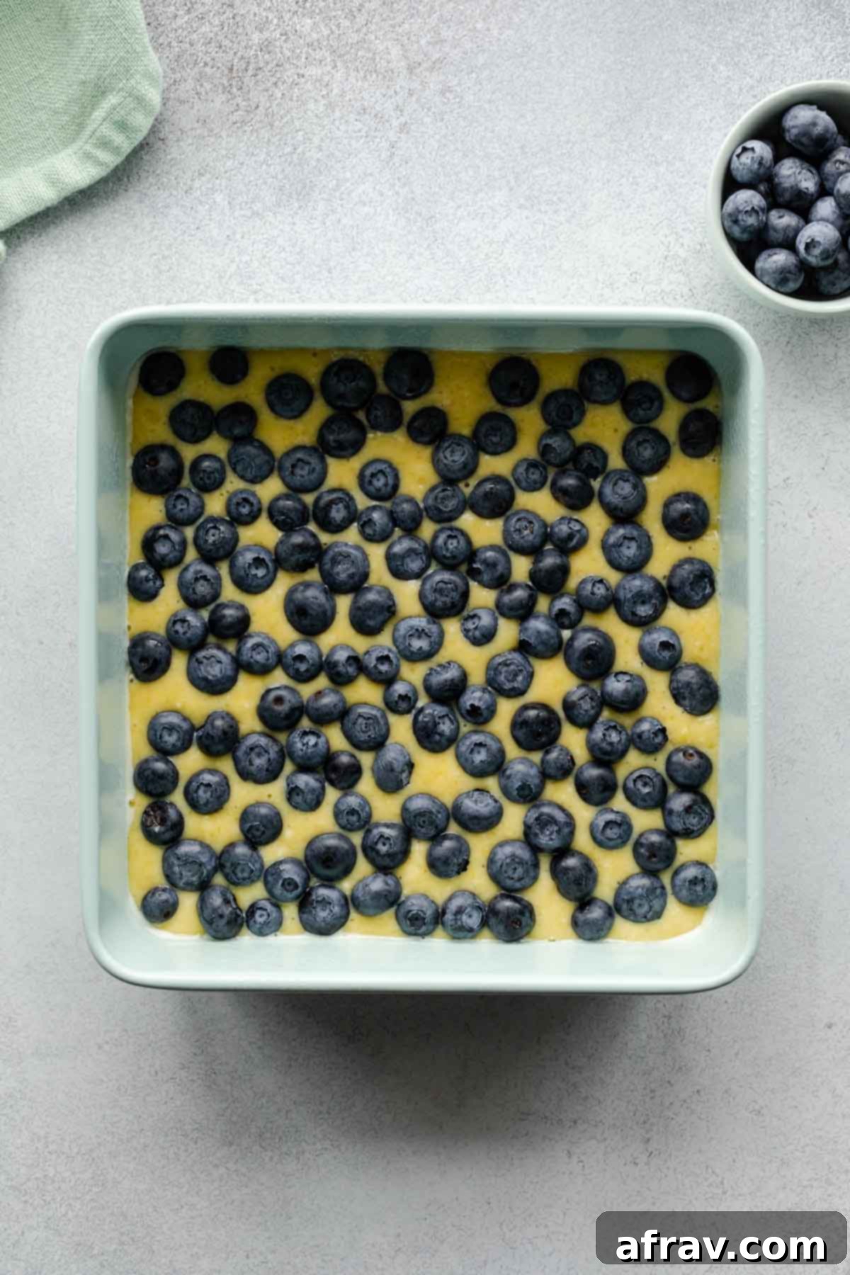 Cake batter in a square pan with blueberries artfully arranged on top, ready for baking.