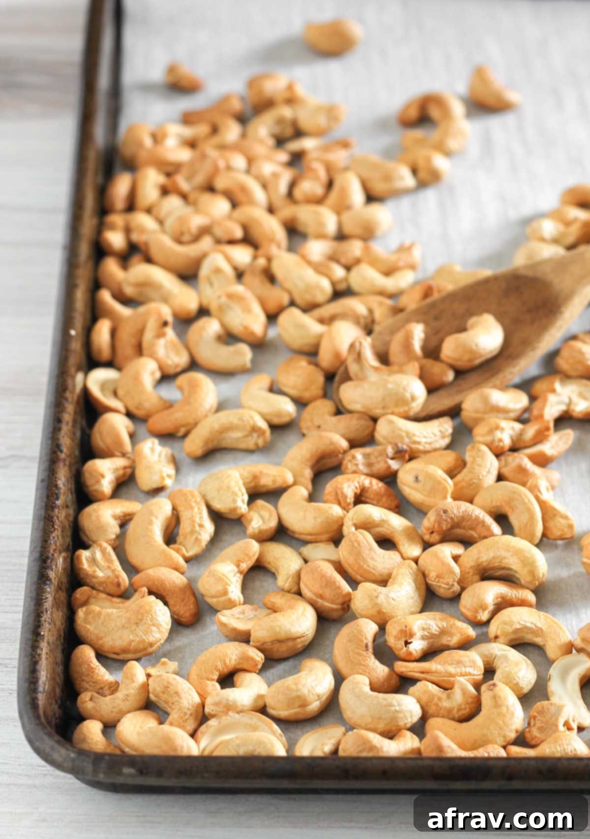 A wooden spoon delicately scooping up a handful of freshly roasted cashews from a vintage baking sheet, highlighting their perfect golden finish.