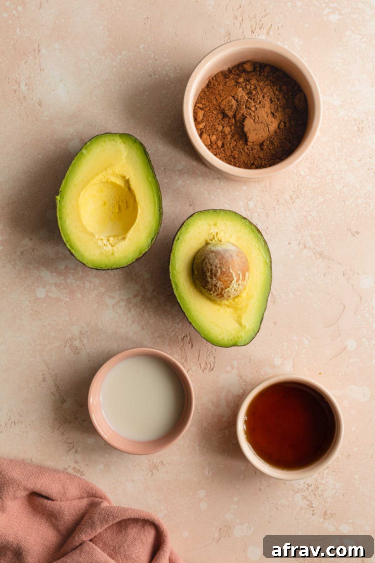Individually arranged ingredients for chocolate avocado frosting: a perfectly ripe avocado, a small bowl of dark cocoa powder, a measuring cup of golden maple syrup, and a splash of almond milk, ready for blending.
