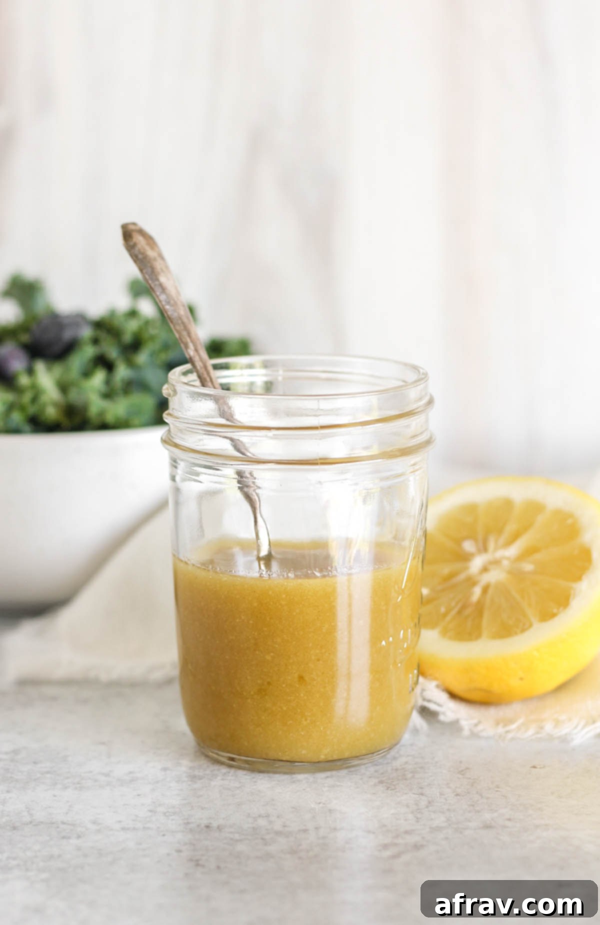 Maple Dijon dressing and a spoon in mason jar with a bowl of greens and a sliced lemon on the background.