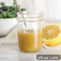Maple Dijon dressing and a spoon in mason jar with a bowl of greens and a sliced lemon on the background.