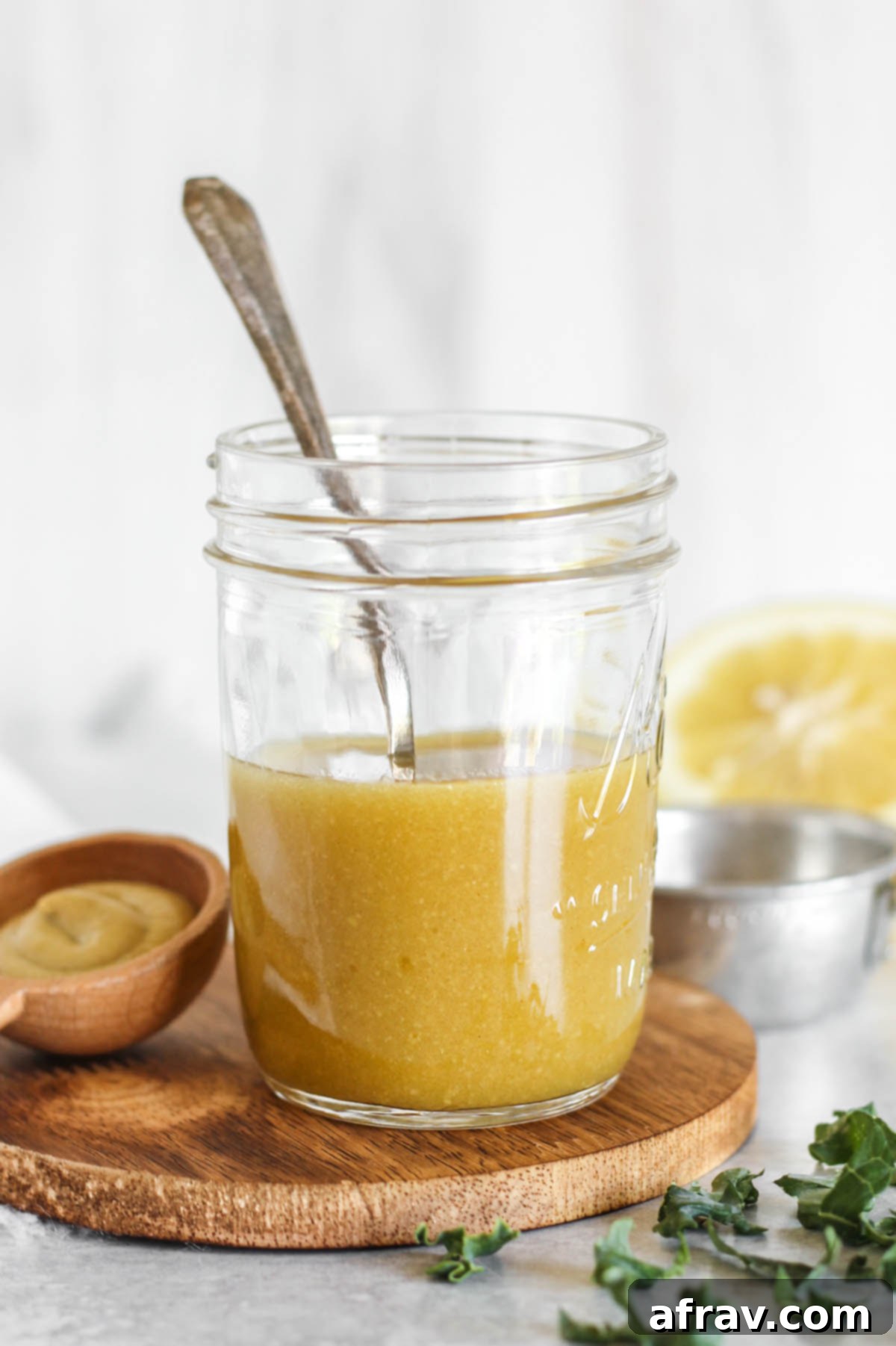Mason jar filled with maple Dijon vinaigrette sitting on a wood tray with greens sprinkled around a lemon on the background.