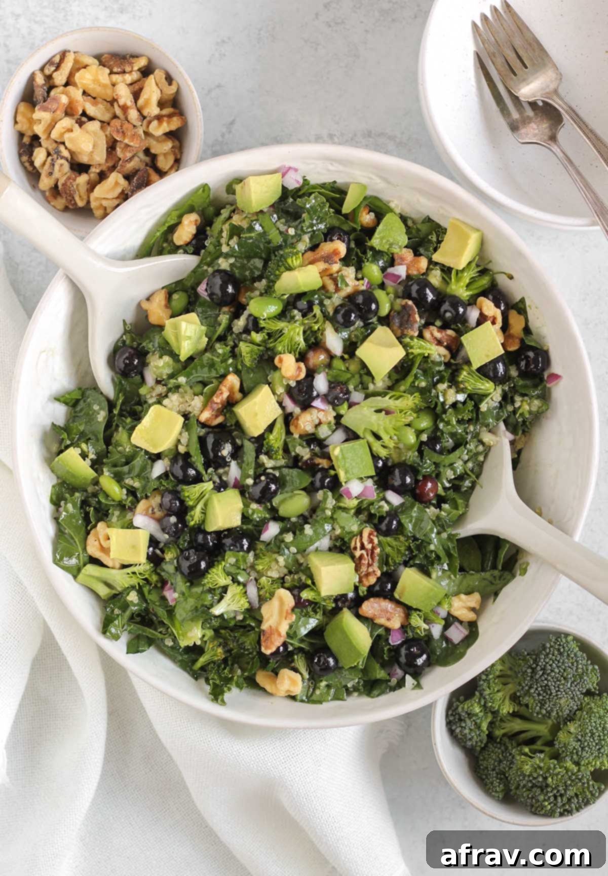 A vibrant superfood salad featuring blueberries, crisp broccoli florets, crunchy walnuts, and creamy avocado, served in a pristine white bowl with elegant white salad utensils. Bowls of fresh broccoli and walnuts are artfully arranged nearby, hinting at the wholesome ingredients within.