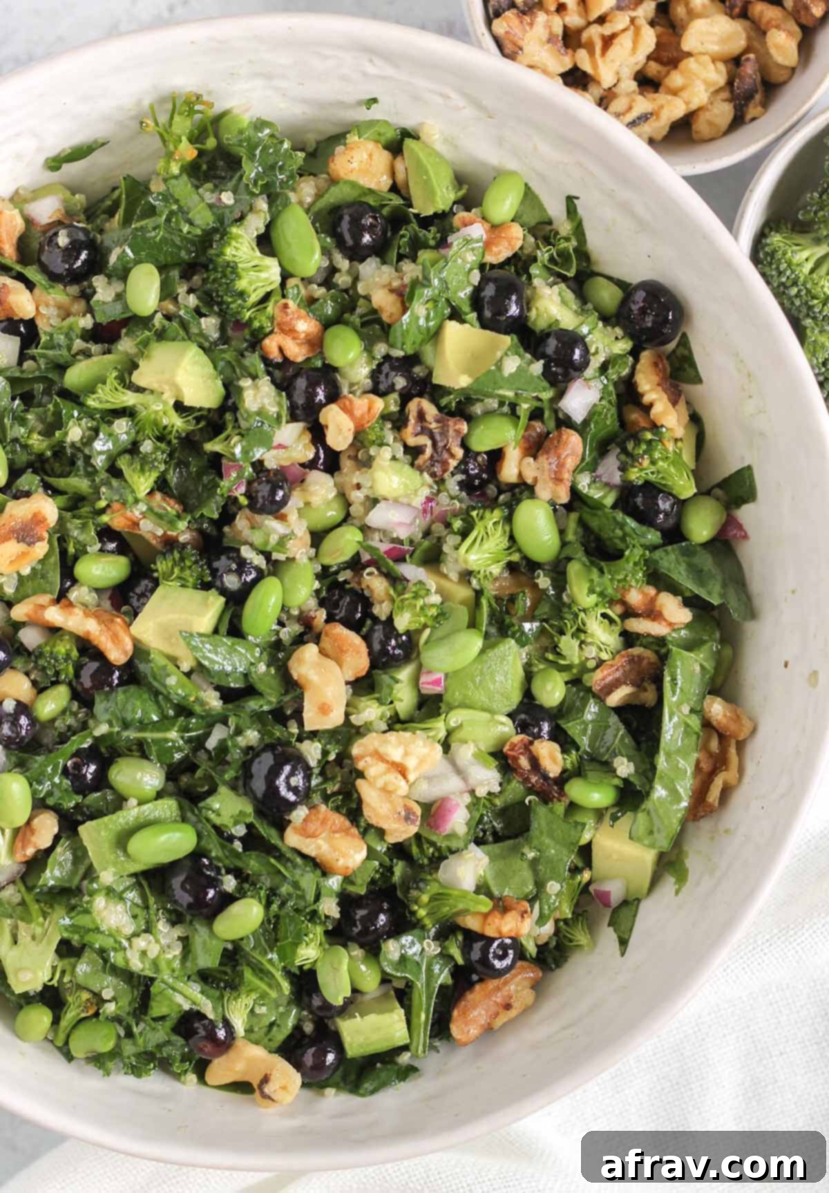 A generous serving of superfood salad elegantly presented in a large white serving bowl, with a small bowl of walnuts and fresh broccoli florets artfully placed on the side, highlighting the wholesome ingredients.