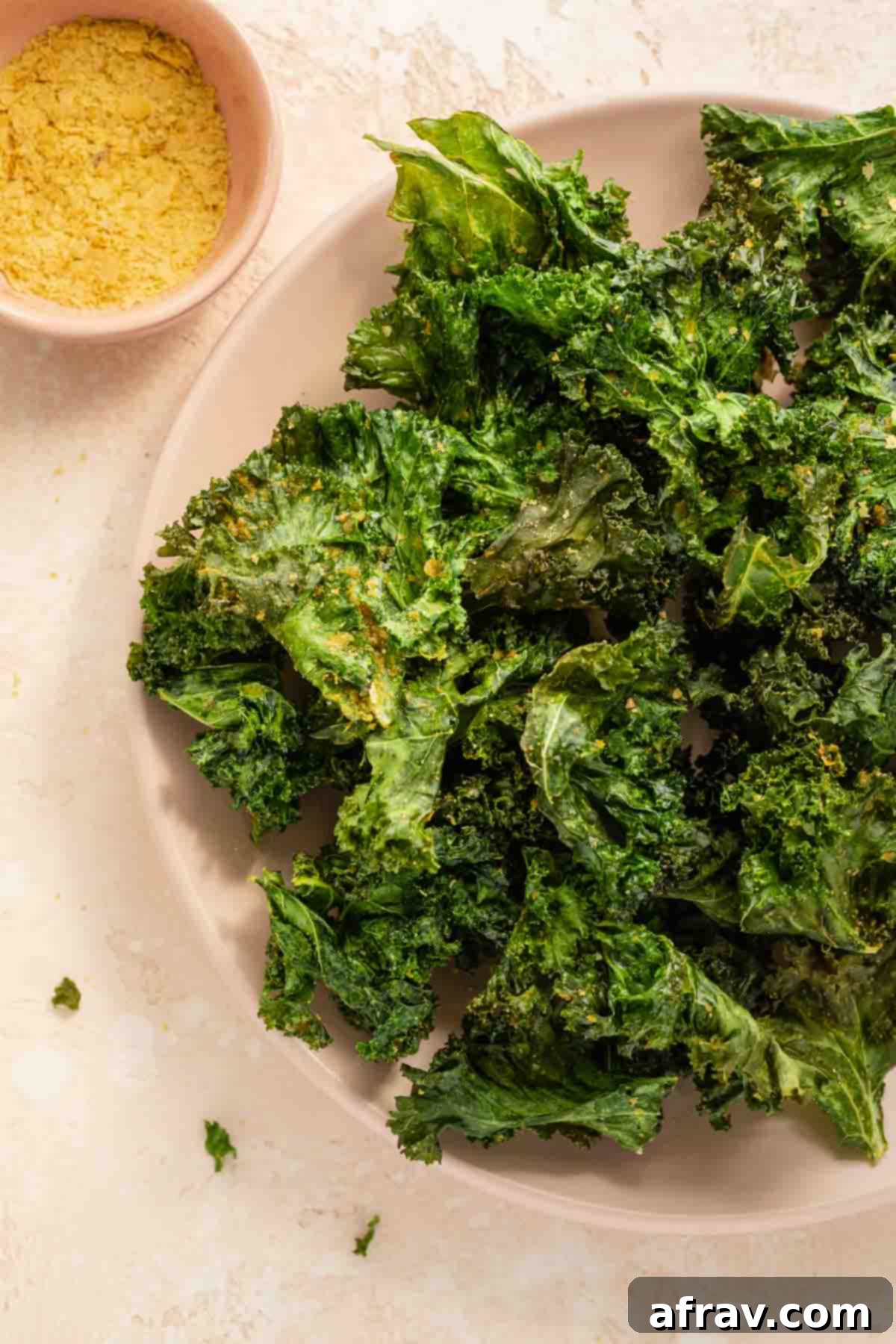 Close-up view of a small pile of golden-green air-fried kale chips on a light pink plate, highlighting their delicate, textured crispness and inviting appearance.