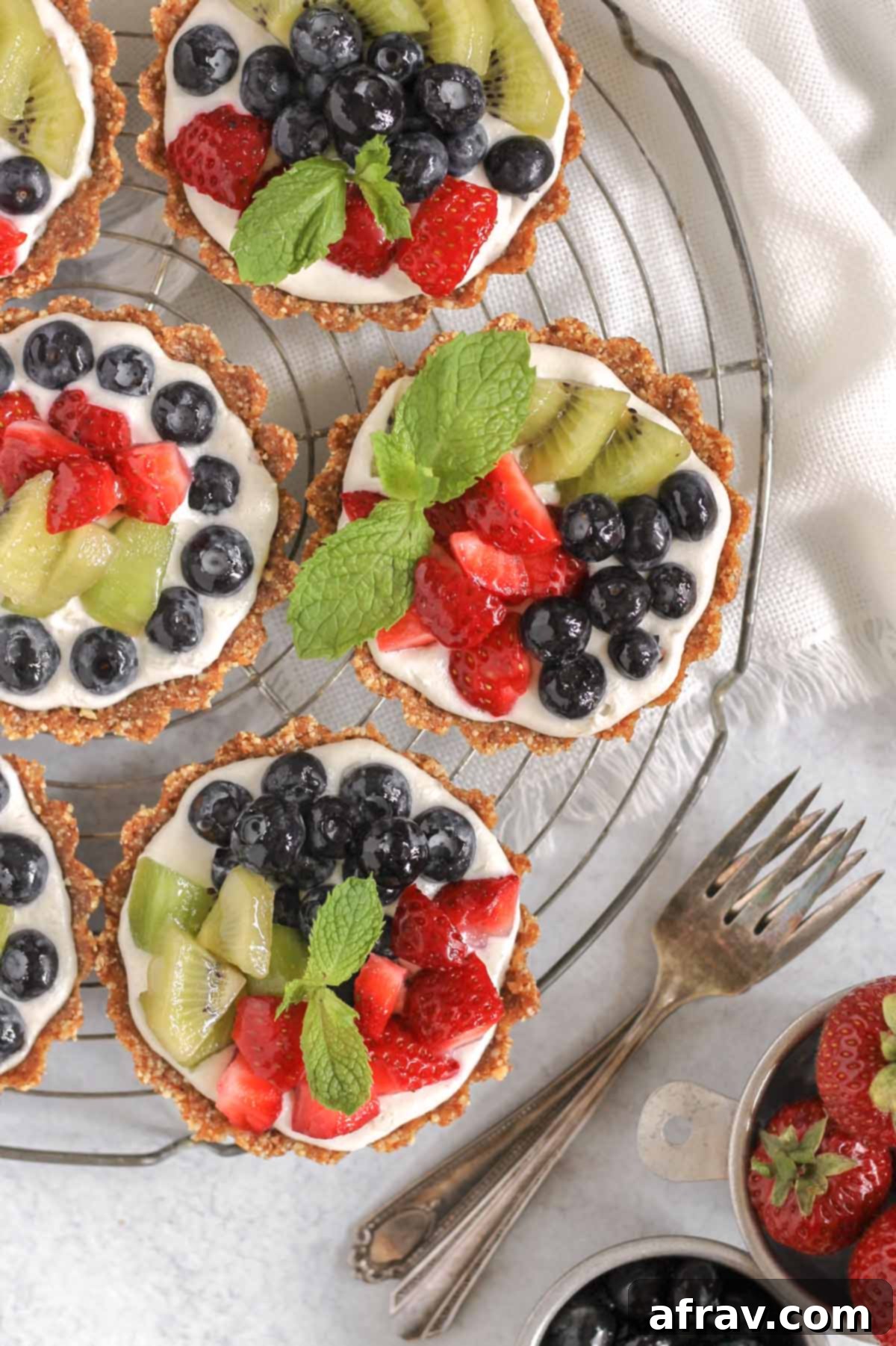 Effortless Fresh Vegan Fruit Tarts 2 Mini vegan fruit tarts artfully arranged on a round wire cooling rack, with two vintage forks resting beside them, ready to be enjoyed.