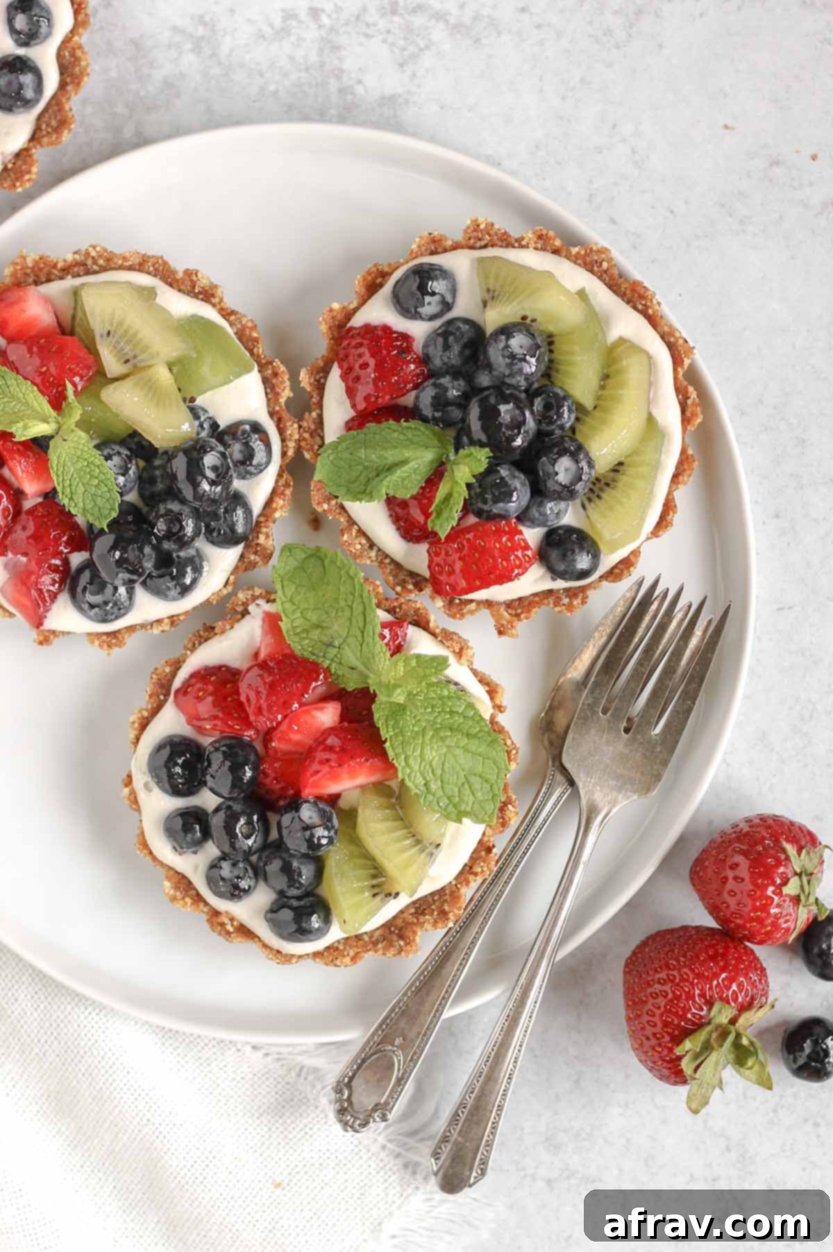 Effortless Fresh Vegan Fruit Tarts 11 Three perfectly plated mini fruit tarts on a small white plate, accompanied by two vintage forks and a scattering of fresh berries off to the side, creating an inviting dessert scene.