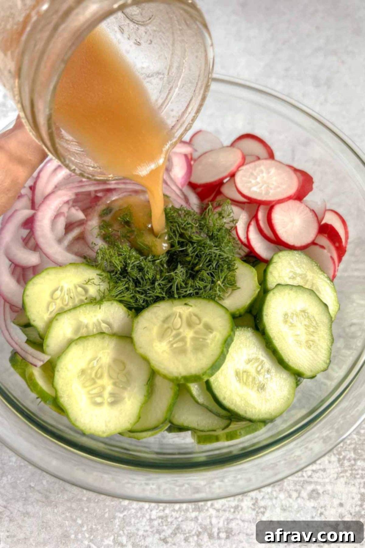 Vinaigrette being poured over cucumber radish salad ingredients in a glass mixing bowl.