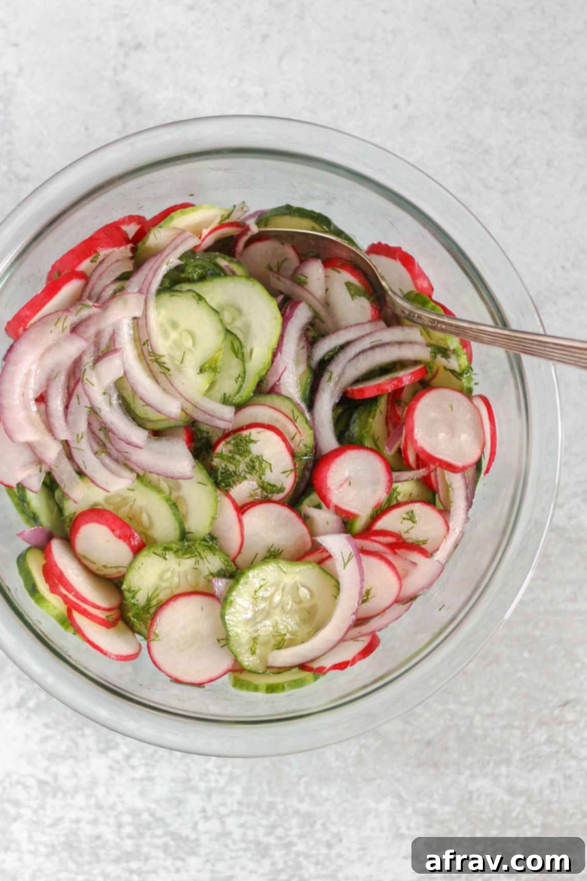 Cucumber, radish, and red onion being mixed in a glass mixing bowl.