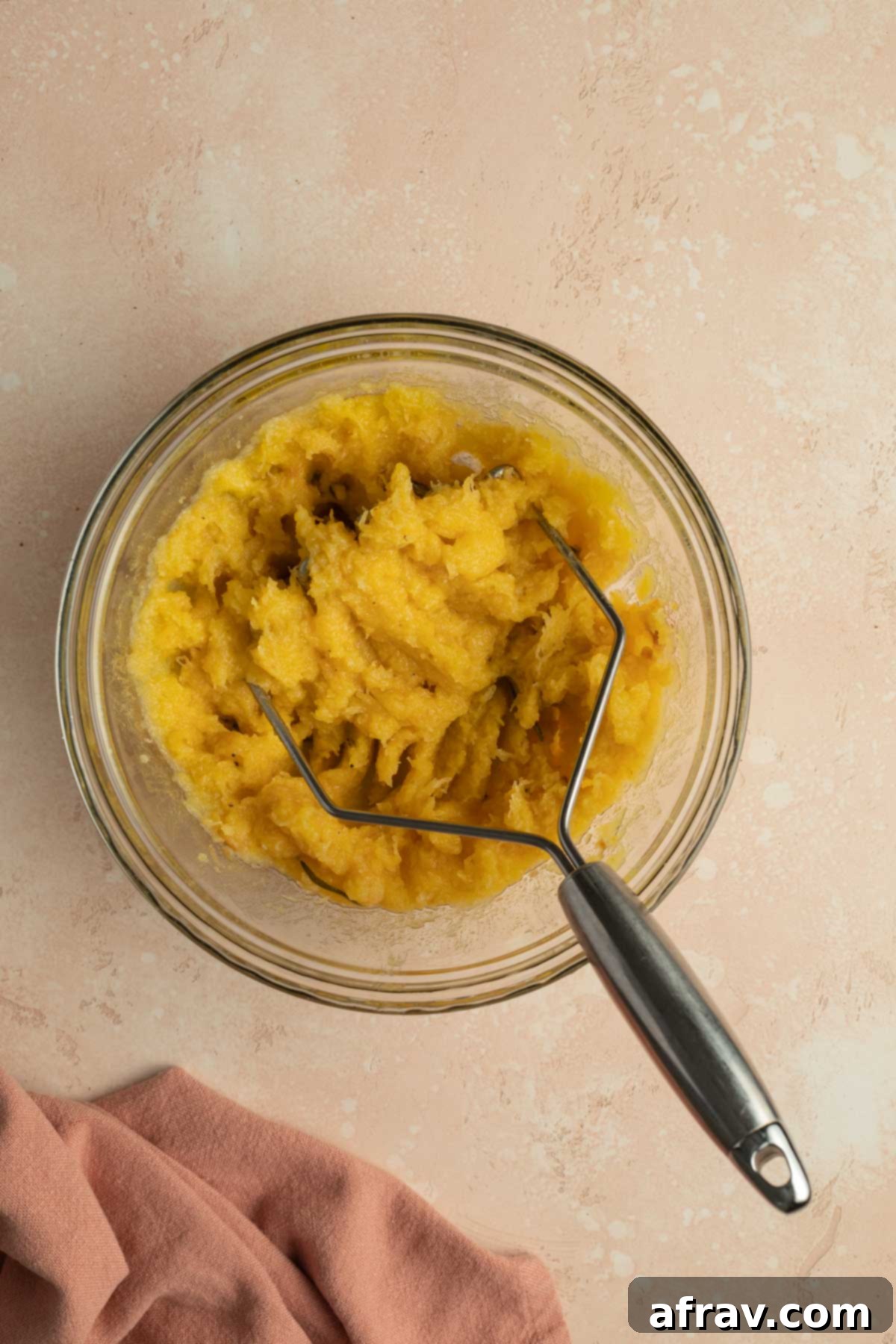 Roasted squash being mashed in a glass bowl with a potato masher.