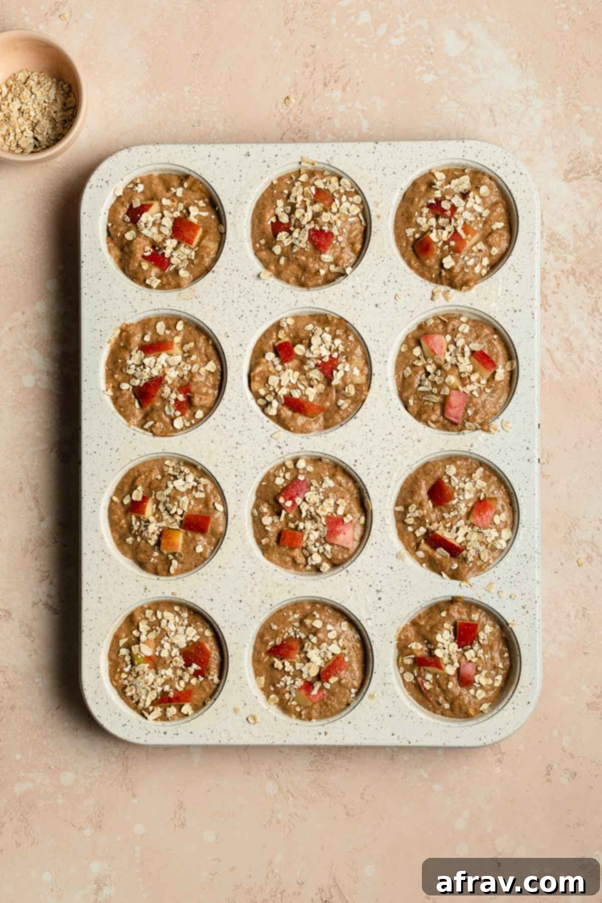Muffin batter scooped into a muffin pan and topped with diced apple and oat flakes, prior to baking.
